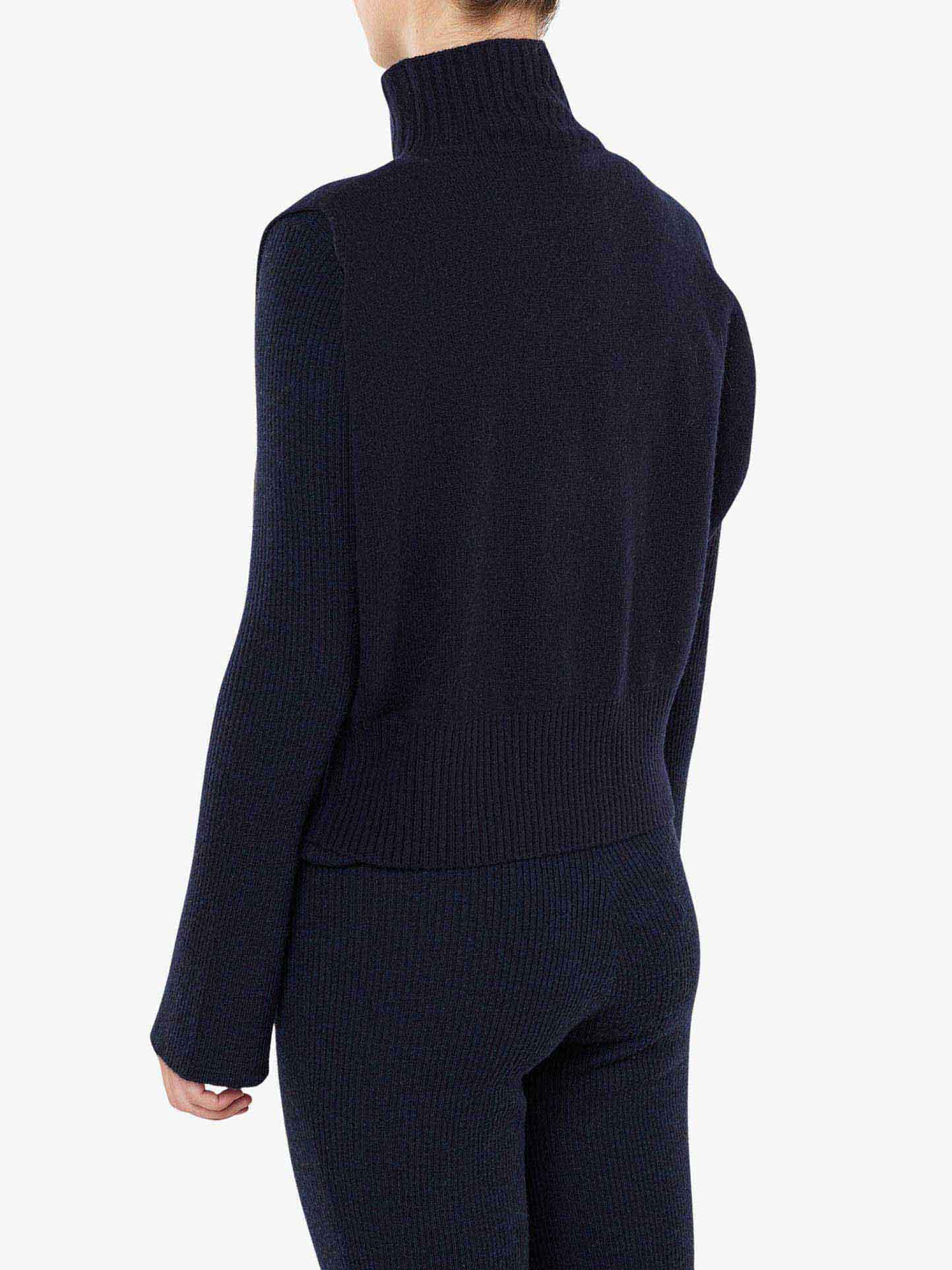 A person is shown from the back wearing a black, long-sleeve, ribbed funnel neck sweater and matching black pants. The sweater has a ribbed texture and is crafted from RWS Extra Fine Merino Wool. The person’s hair is tied back, and the background is plain white. They are also wearing the Geilo Vest Women by We Norwegians.