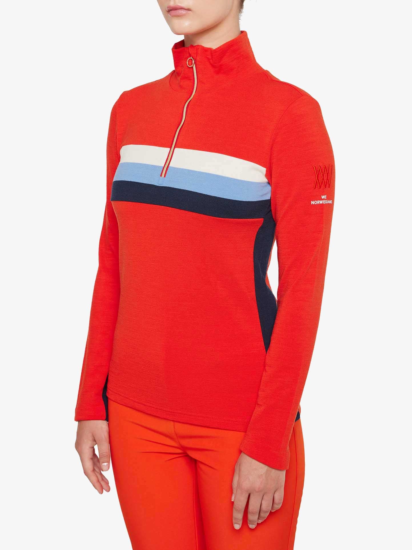 A person wearing an orange We Norwegians Ski 1/4-Zip Women long-sleeve pullover with a half-zip and horizontal white and blue stripes on the chest and left shoulder. The Merino wool pullover features black side panels, a logo on the left sleeve, and is paired with matching orange pants.