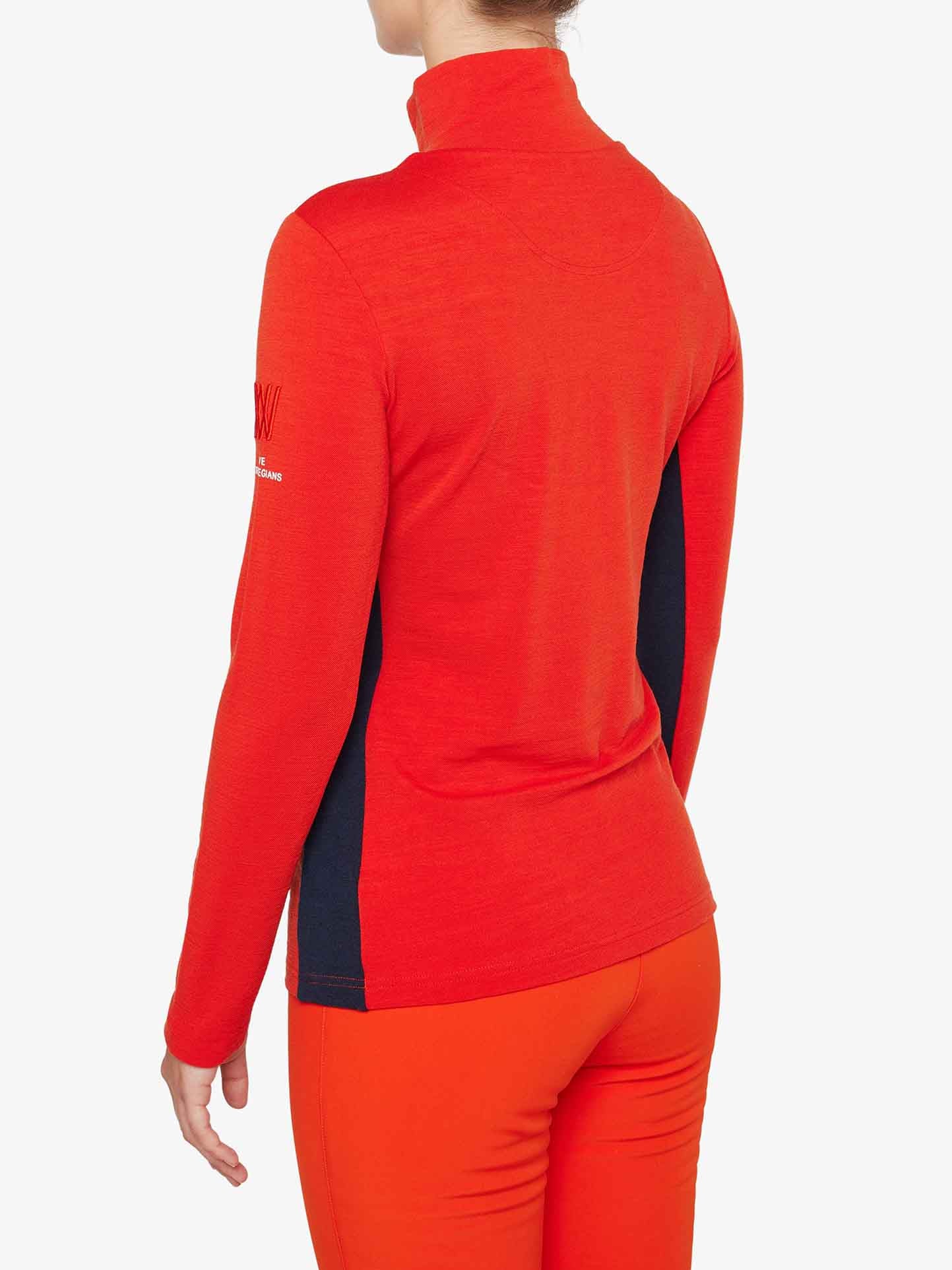 A person is wearing a long-sleeved, bright red Ski 1/4-Zip Women shirt from We Norwegians with black side panels. The shirt features a high collar and visible stitching on the back and sleeves. Made of Merino wool, it serves as an excellent base layer. The person is also wearing matching red pants against a plain white background.