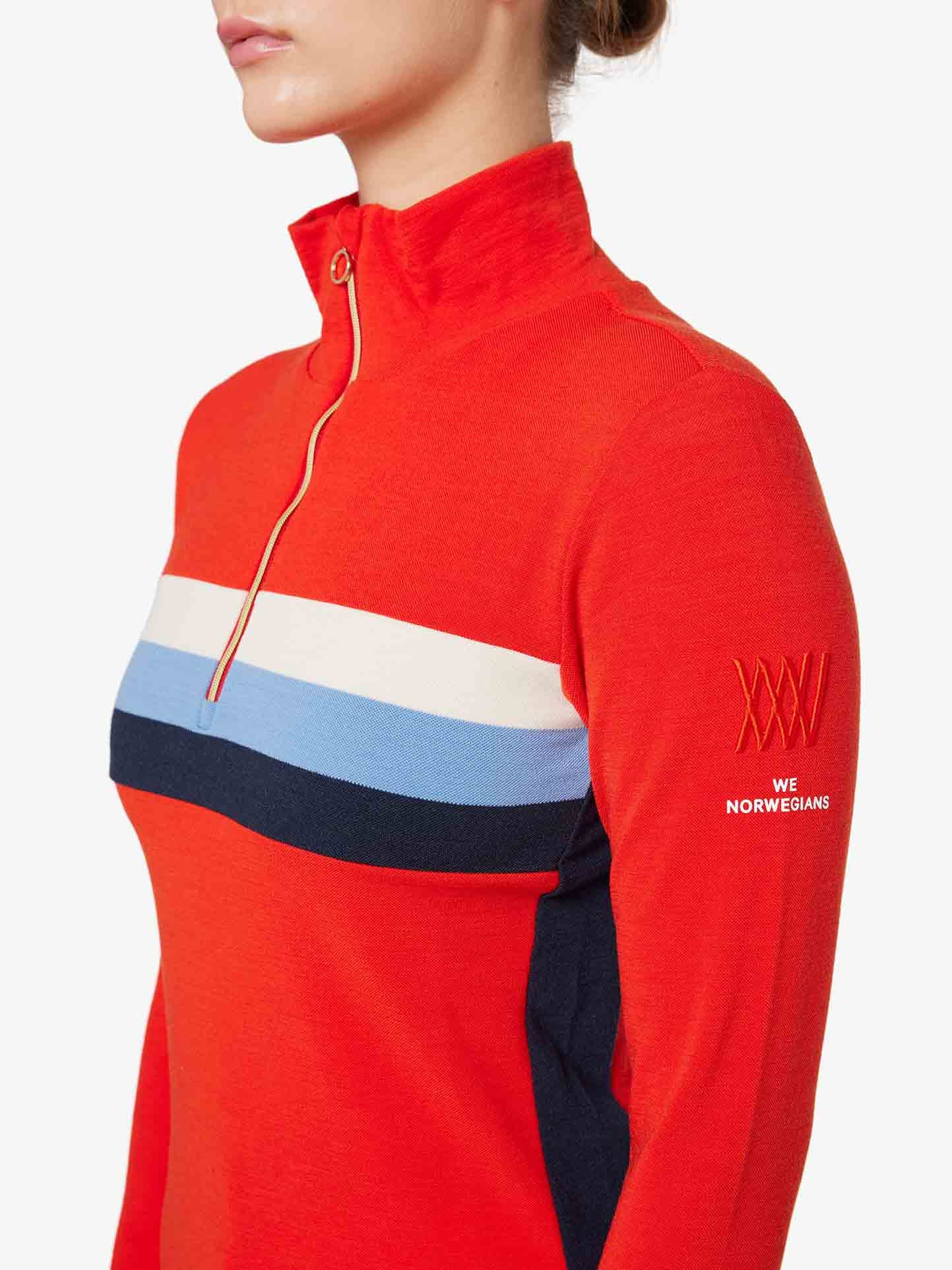 A person wearing a vibrant orange Ski 1/4-Zip Women sweater by We Norwegians, showcasing horizontal white, blue, and navy stripes across the chest. Made from Merino wool, the sleeve is adorned with the text "XXX" and "We Norwegians." The individual has their hair tied back and is photographed from the neck down.