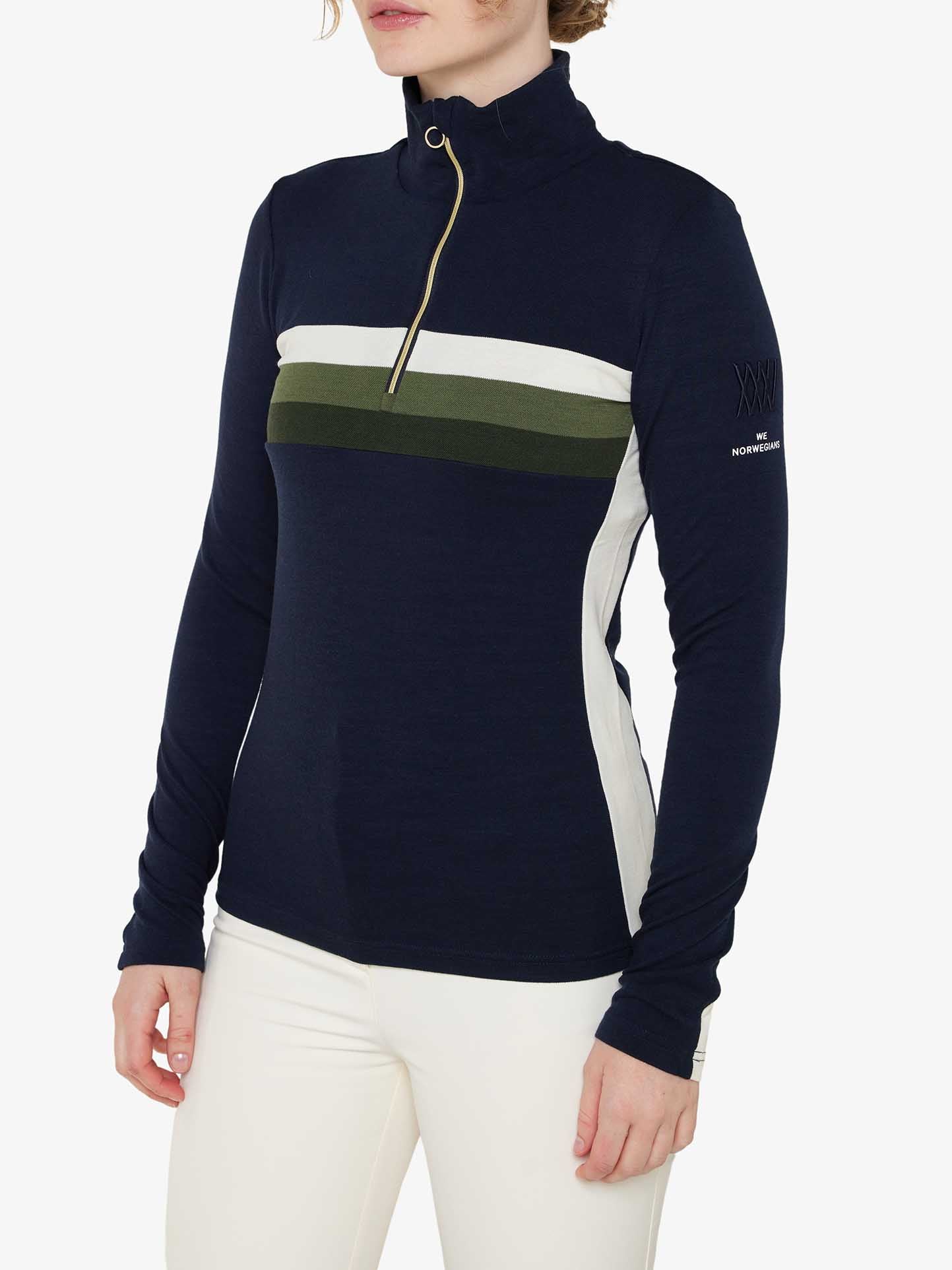 A person wearing a navy blue We Norwegians Ski 1/4-Zip Women long-sleeve top with a high collar, zip neck, and horizontal stripes in white and olive green across the chest. The person is also wearing white pants and is standing against a plain white background.