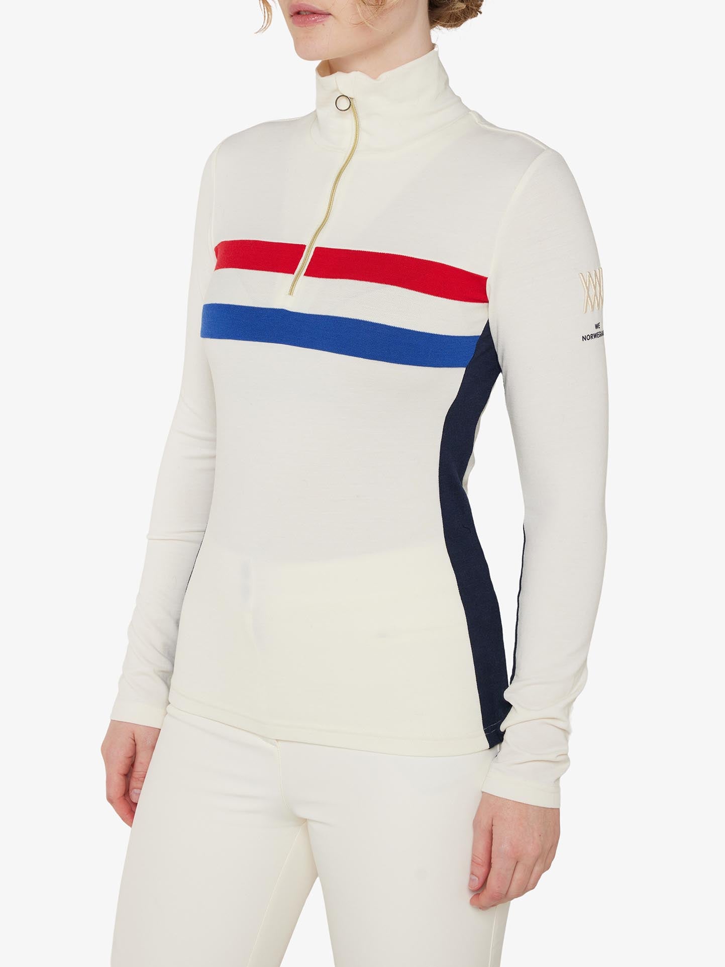 A person is wearing a white long-sleeve, high-neck We Norwegians Ski 1/4-Zip Women top with red, white, and blue horizontal stripes across the chest. The top features a half-zipper and dark blue side panels and is made from Merino wool. They're also sporting matching white pants.