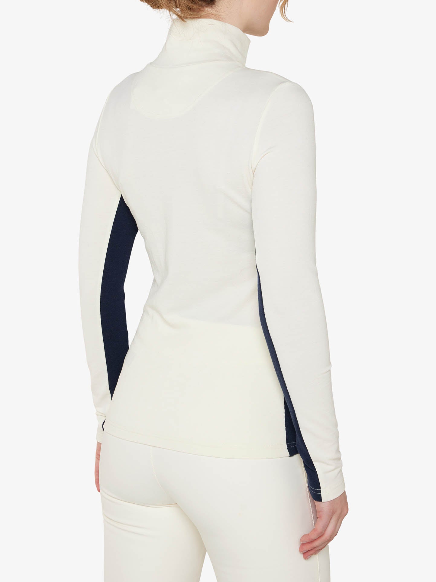 A person wearing a white Ski 1/4-Zip Women by We Norwegians stands with their back to the camera. The Merino wool shirt features navy blue side panels and is paired with matching white pants. The background is plain and white.