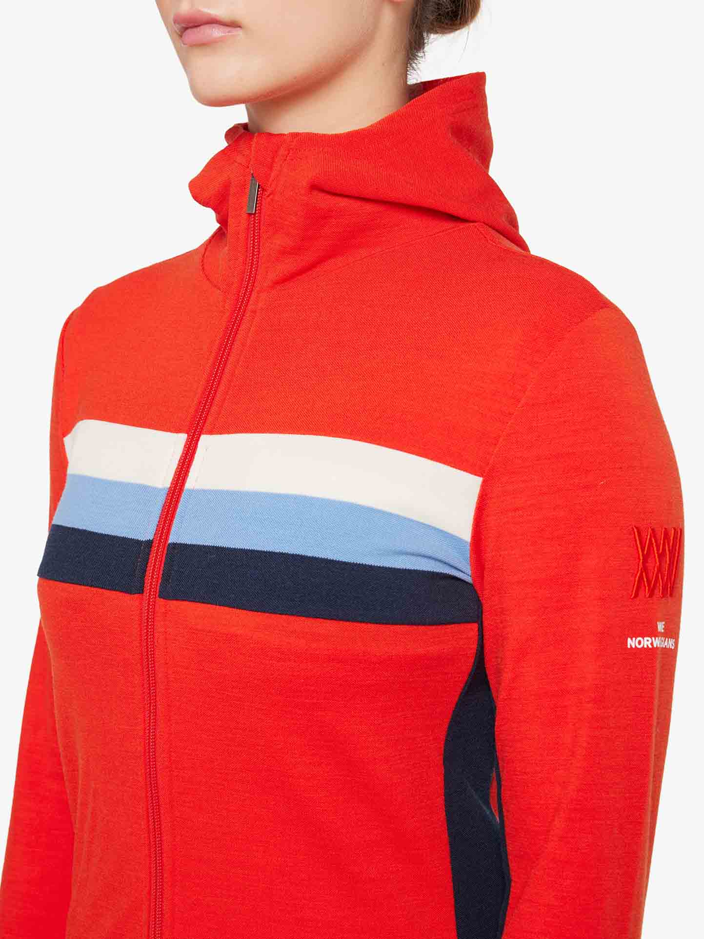 A person wears the We Norwegians Ski Fullzip Hoodie for women, featuring retro color block panels in white, blue, and navy stripes across the chest. The red jacket includes a hood and has a design on the left sleeve with "XXX" and "NORWAY" in small text. The person's head is partially visible.