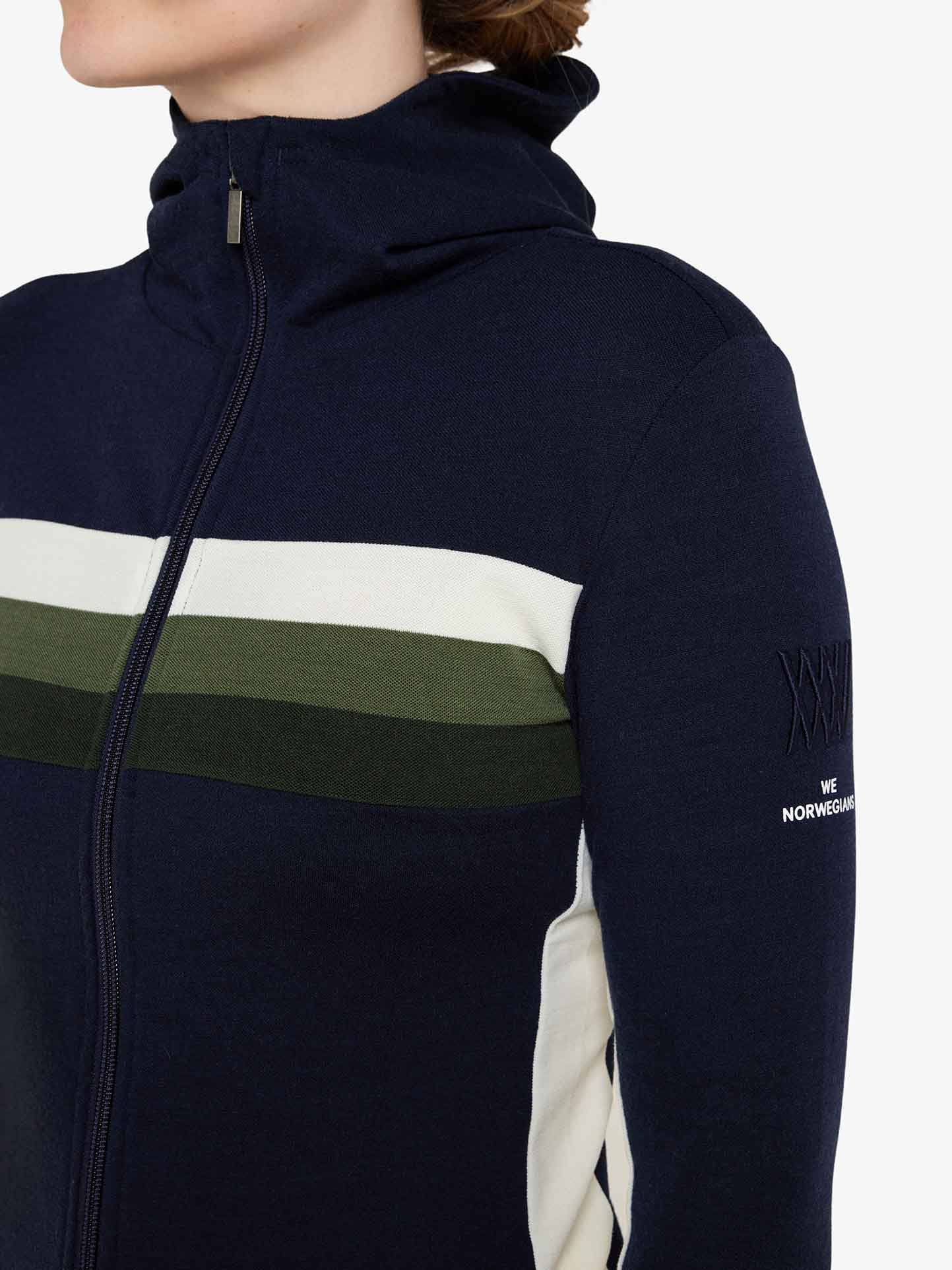 A close-up image of a person wearing a dark navy Ski Fullzip Hoodie Women by We Norwegians, featuring horizontal white and green stripes across the chest, retro color block panels, and subtle embroidered text on the sleeve that reads "XXXIX WE NORWEGIANS.