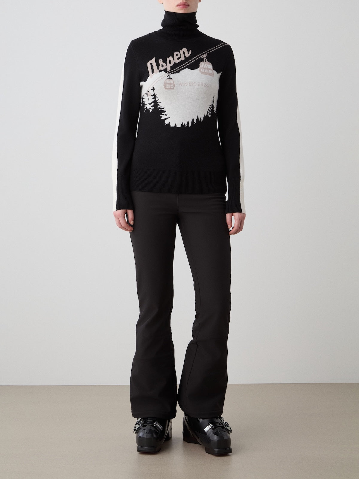 A person wears the We Norwegians Afterski Sweater Women in black and white, featuring an “Aspen” mountain design, styled with black fitted pants and black platform shoes, standing against a plain light background.