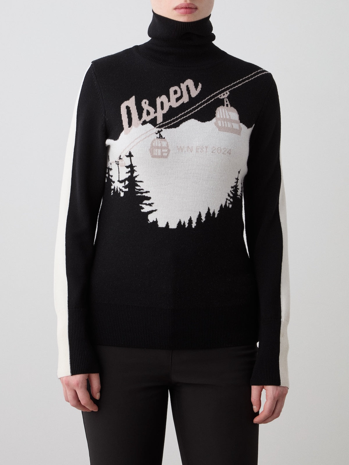 A woman wears the We Norwegians Afterski Sweater, featuring “Aspen,” a cable car motif, white sleeves, and winter landscape artwork. This cozy après-ski piece combines style and comfort for chilly days.