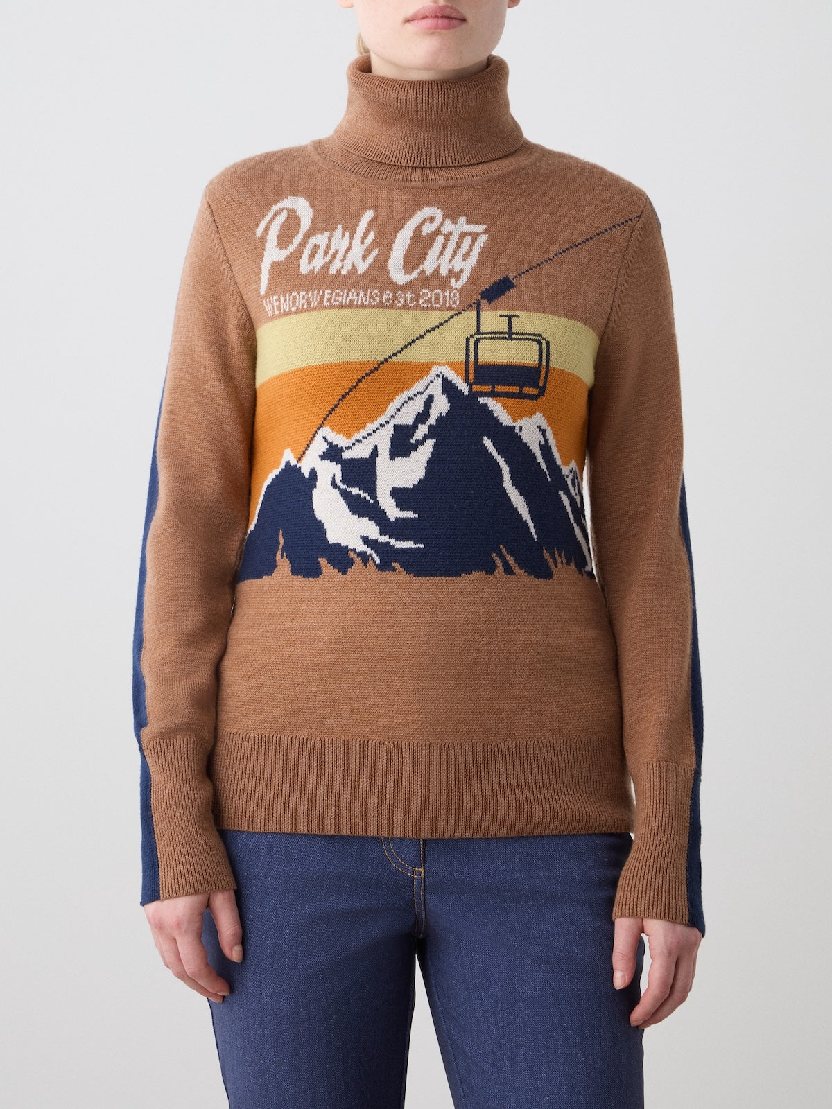 A woman wears the We Norwegians Afterski Sweater in brown, featuring "Park City" and "Memories & Dreams Est. 2018" text with a mountain, cable car, and sunset design, styled with blue jeans.