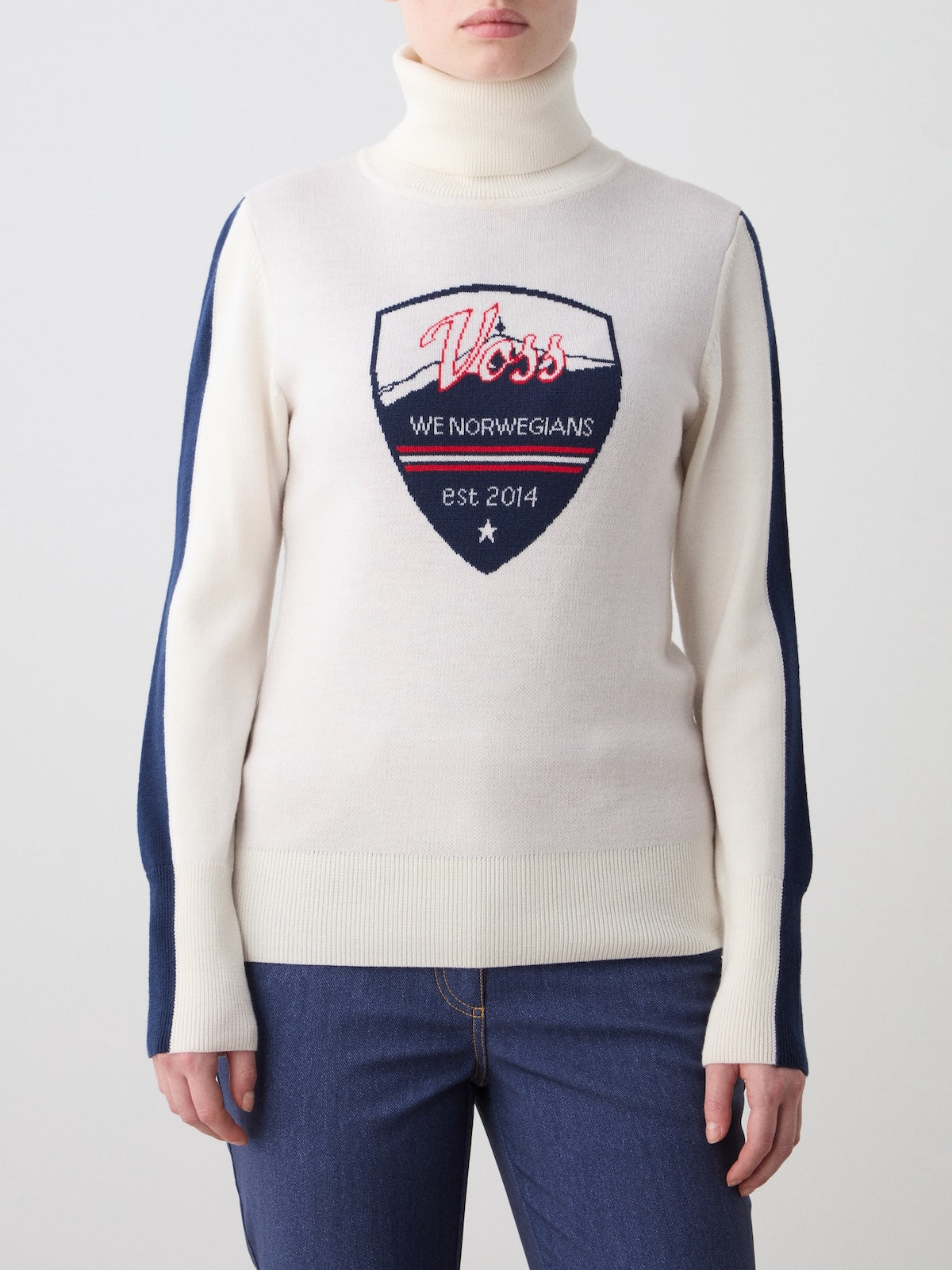 A person is wearing the We Norwegians Afterski Sweater Women, a cream retro ski sweater with blue sleeves and a chest shield reading "Voss, We Norwegians, est 2014," styled with blue jeans for a classic après-ski winter look.