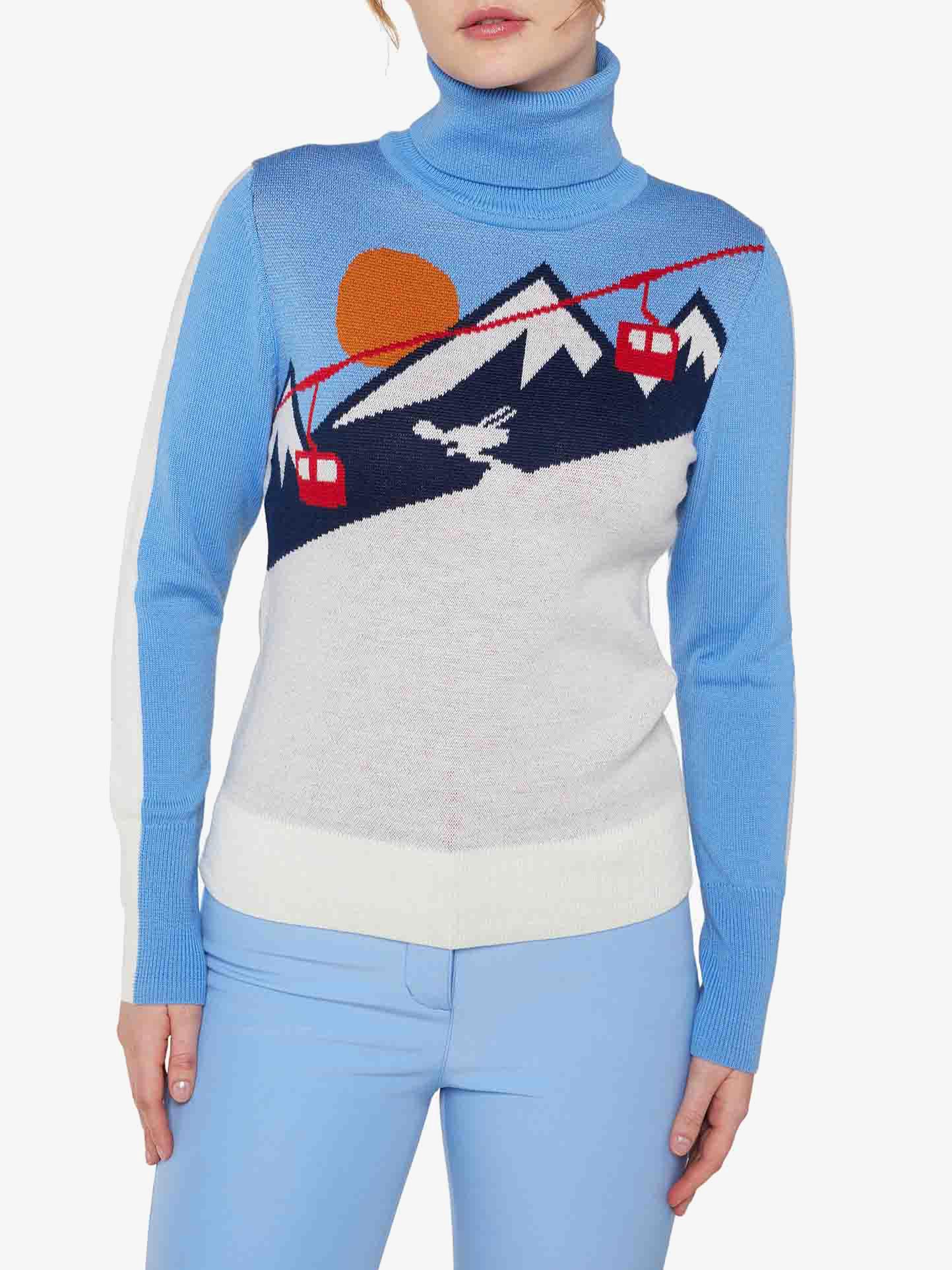 A woman wearing the We Norwegians' light blue "Afterski Sweater Women" made of merino wool, featuring a mountain scene with cable cars, a bird, and a setting sun design on the front. She is also dressed in matching light blue pants against a plain white background.