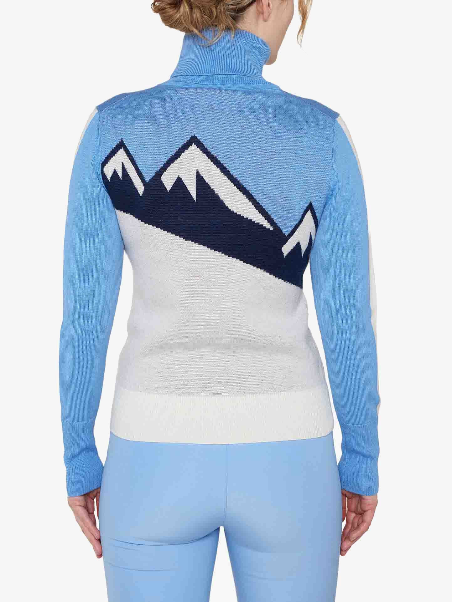 A person wearing the We Norwegians Afterski Sweater Women, a light blue and white merino wool turtleneck sweater featuring dark blue mountain peaks on the back. The individual is also wearing matching light blue pants, embodying the perfect après-ski look as they face away, showcasing the back of the outfit.