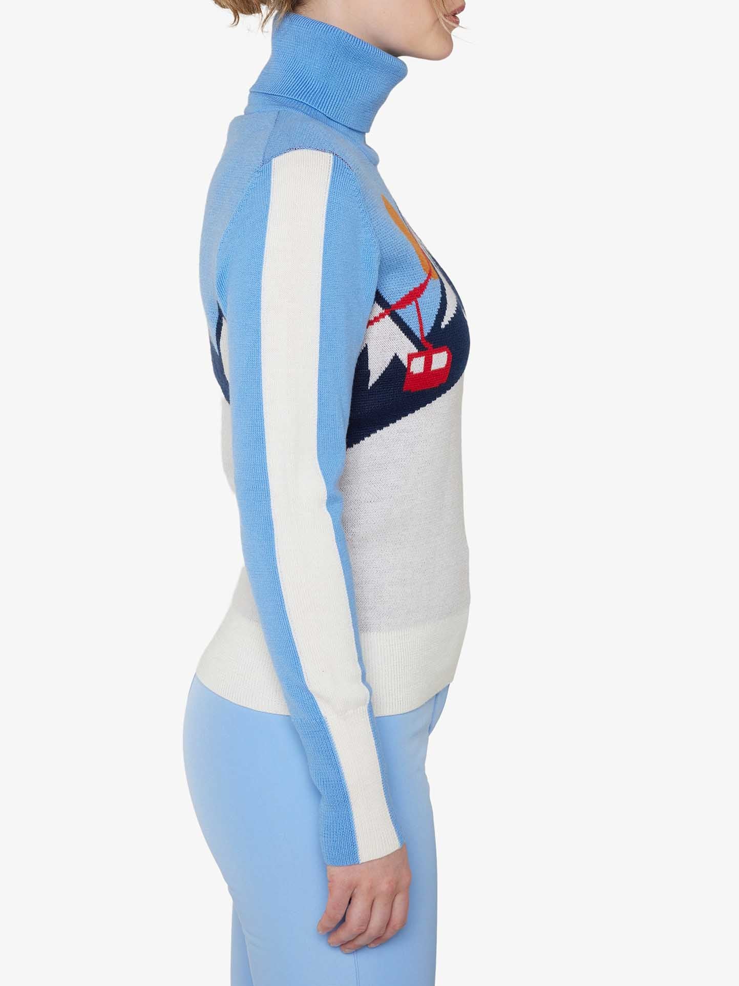 Side profile of a woman wearing the We Norwegians Afterski Sweater Women in light blue and white, crafted from merino wool and adorned with a colorful graphic design on the chest. She is also dressed in matching light blue leggings. The plain white background evokes cozy après-ski evenings.