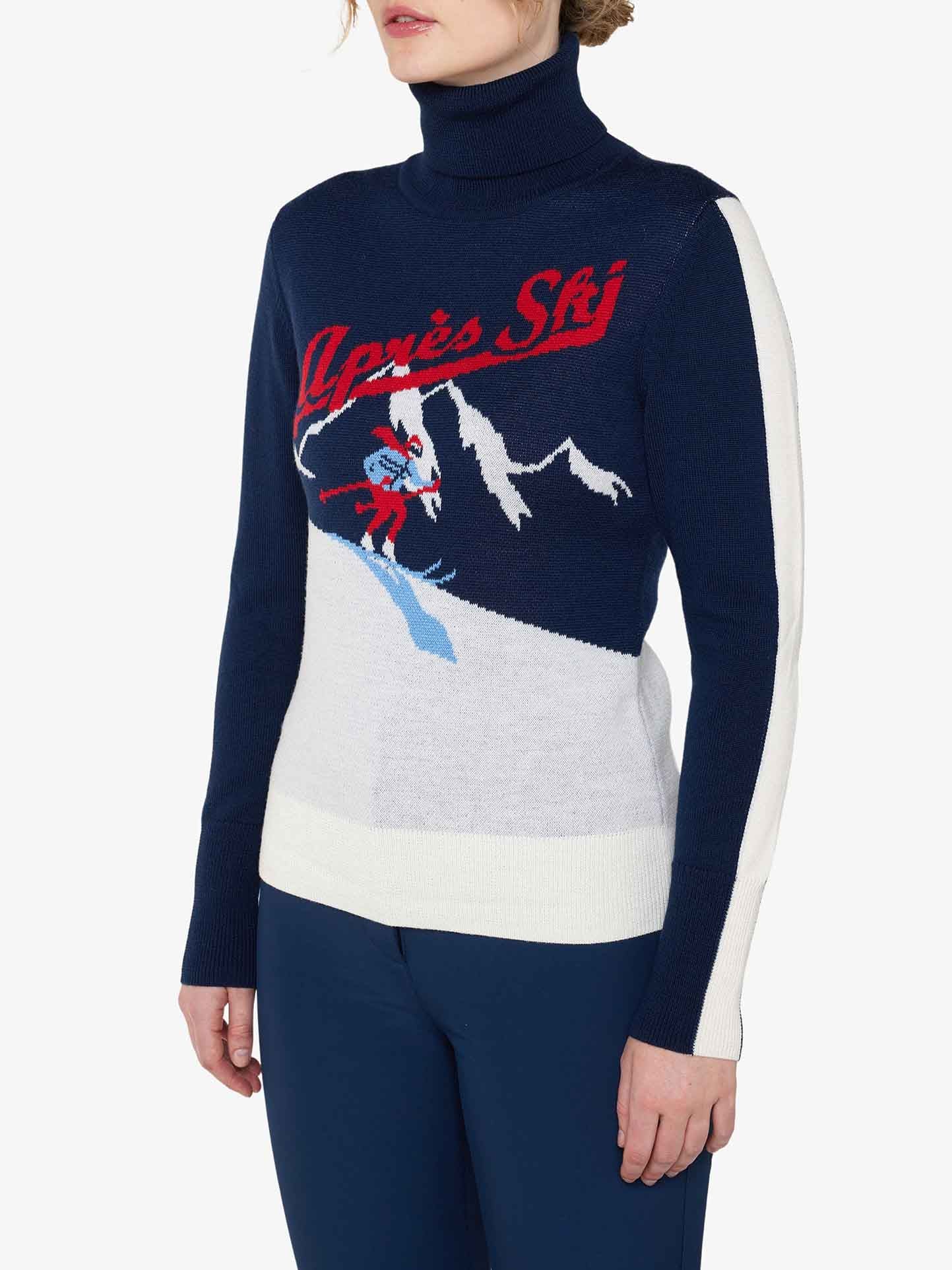 A person wearing the Afterski Sweater Women by We Norwegians, a cozy blue turtleneck made of merino wool, stands against a white background. The sweater features white sleeves and displays white and red graphics of a skiing scene with the text "Après Ski" alongside a snow-covered mountain design in the middle.