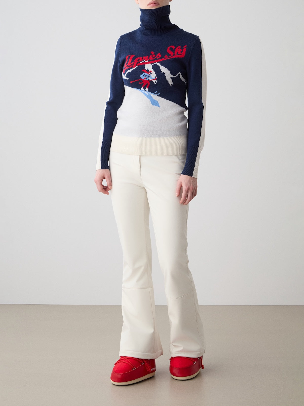 A person stands against a plain background wearing the We Norwegians Afterski Sweater Women with a skiing graphic and “Après Ski” text, crafted from washable merino wool, paired with white pants and bright red shoes. Their face is not visible.