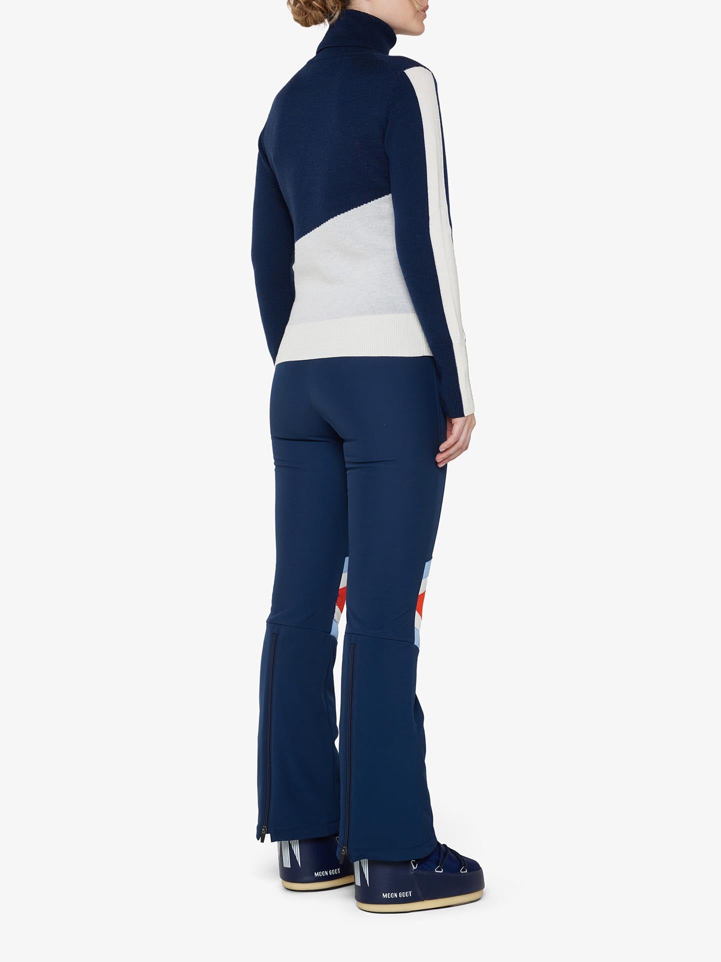 A person is seen from the back, wearing the fitted navy blue and white "Afterski Sweater Women" by We Norwegians, made of merino wool, paired with navy blue ski pants featuring zippered flares at the hem and navy blue ski boots. The attire looks sporty and perfect for winter activities or après-ski relaxation.