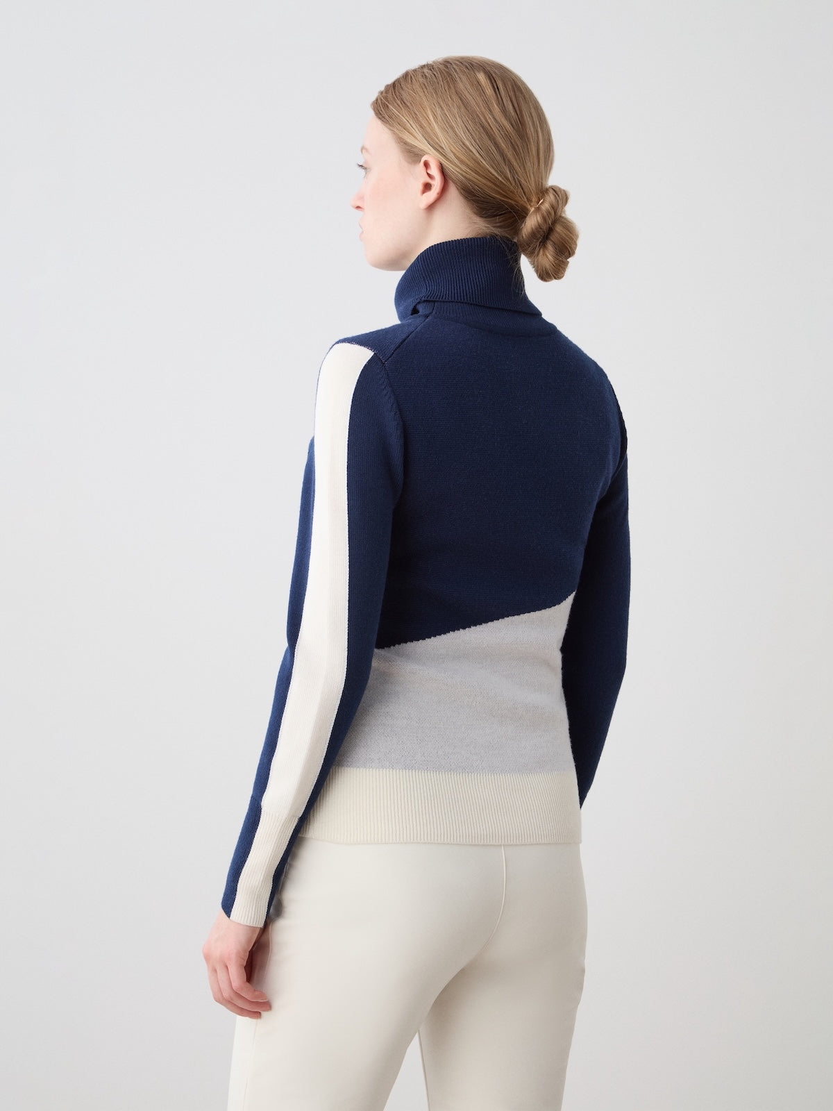 A woman with light brown hair in a low bun, facing away, models the We Norwegians Afterski Sweater Women in navy, cream, and light gray with white pants against a plain light background.