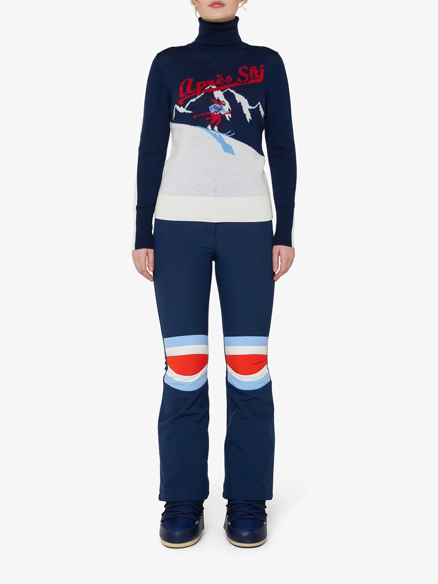 A person is standing against a plain backdrop wearing a black "Afterski Sweater Women" by We Norwegians, made of cozy merino wool and adorned with red lettering and a skiing graphic. They are also sporting blue ski pants featuring white, red, and light blue stripes at the knees, complemented by black ski boots.