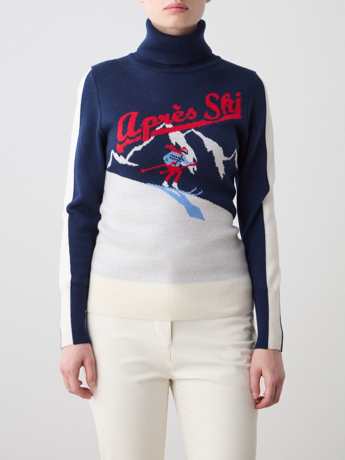 A woman wearing the We Norwegians Afterski Sweater—a blue and white après-ski knit featuring an “Après Ski” slogan and a skier on snowy mountains design—paired with white pants.