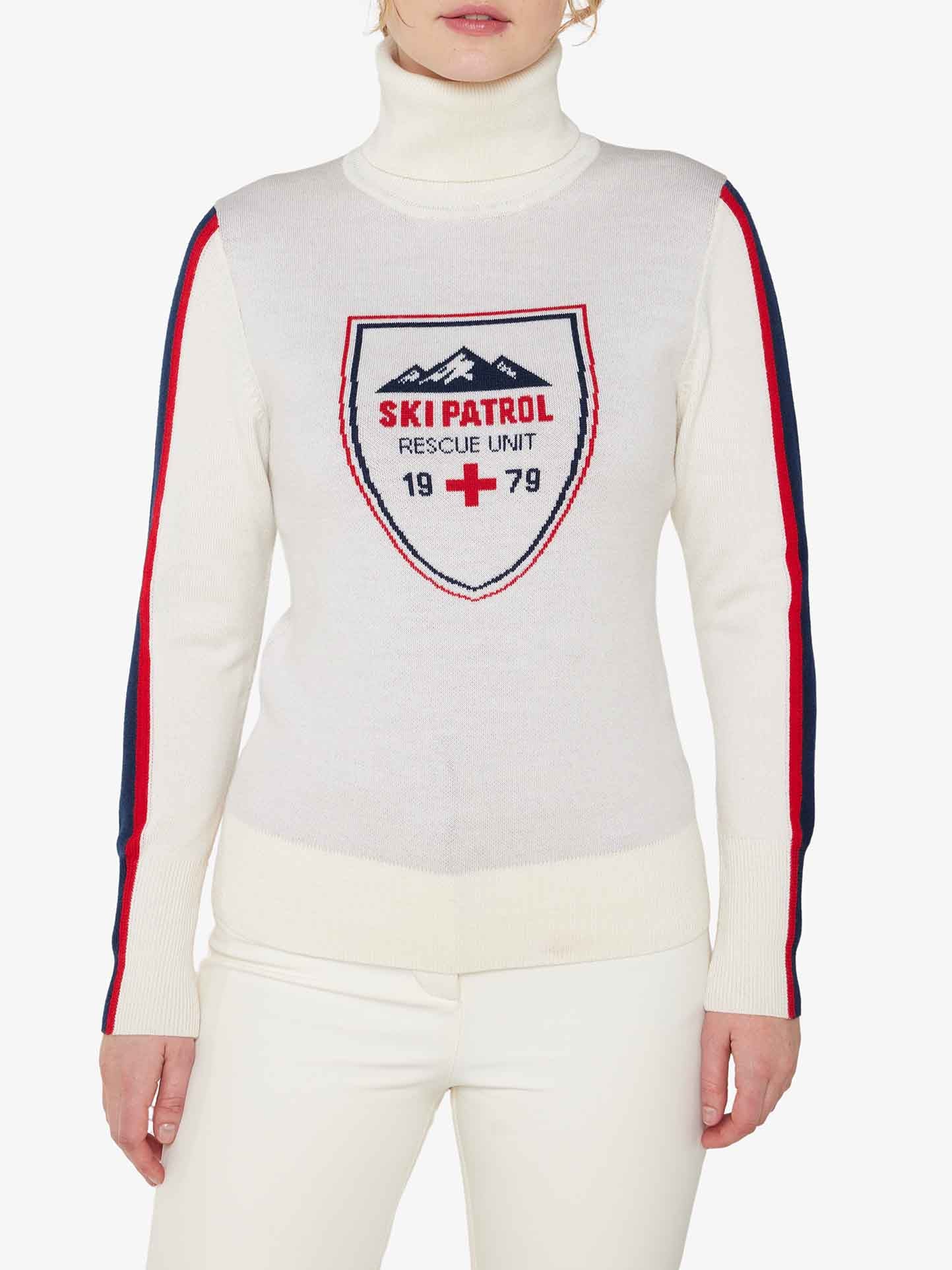 A person wearing the "Afterski Sweater Women" by We Norwegians, featuring a white turtleneck design with red and blue stripes on the sleeves and a "Ski Patrol Rescue Unit 1979" emblem on the chest, reminiscent of classic après ski style. The ensemble includes white pants, set against a plain white background.