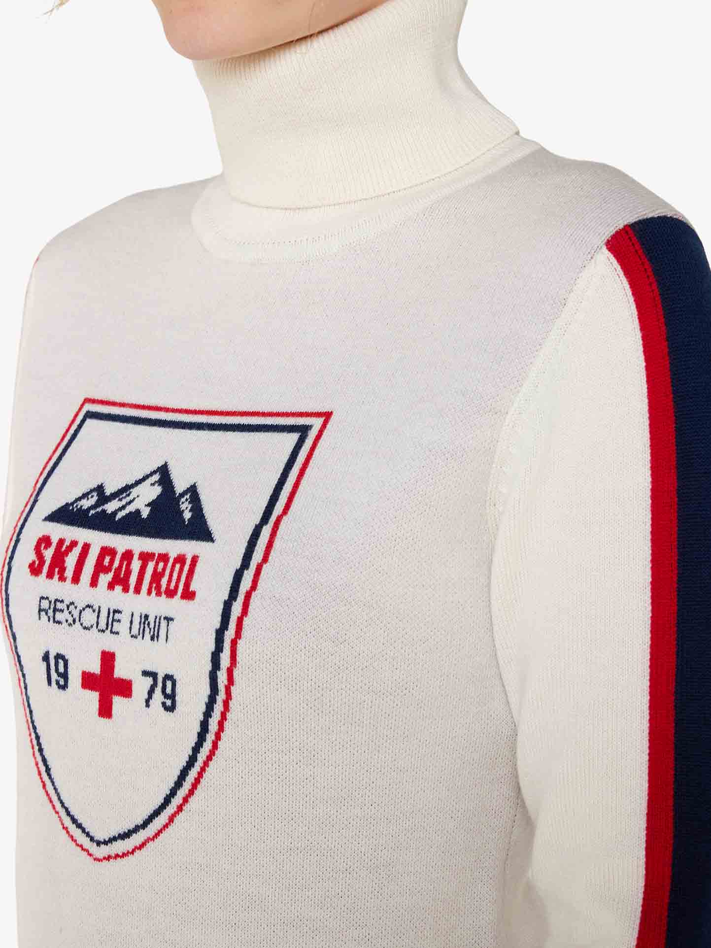 Close-up of a person wearing the Afterski Sweater Women by We Norwegians. The white merino wool turtleneck features a "Ski Patrol Rescue Unit 1979" logo on the chest, complete with a mountain graphic and red cross symbol. Stylish red and blue stripes adorn the sleeves, making it perfect for après-ski gatherings.