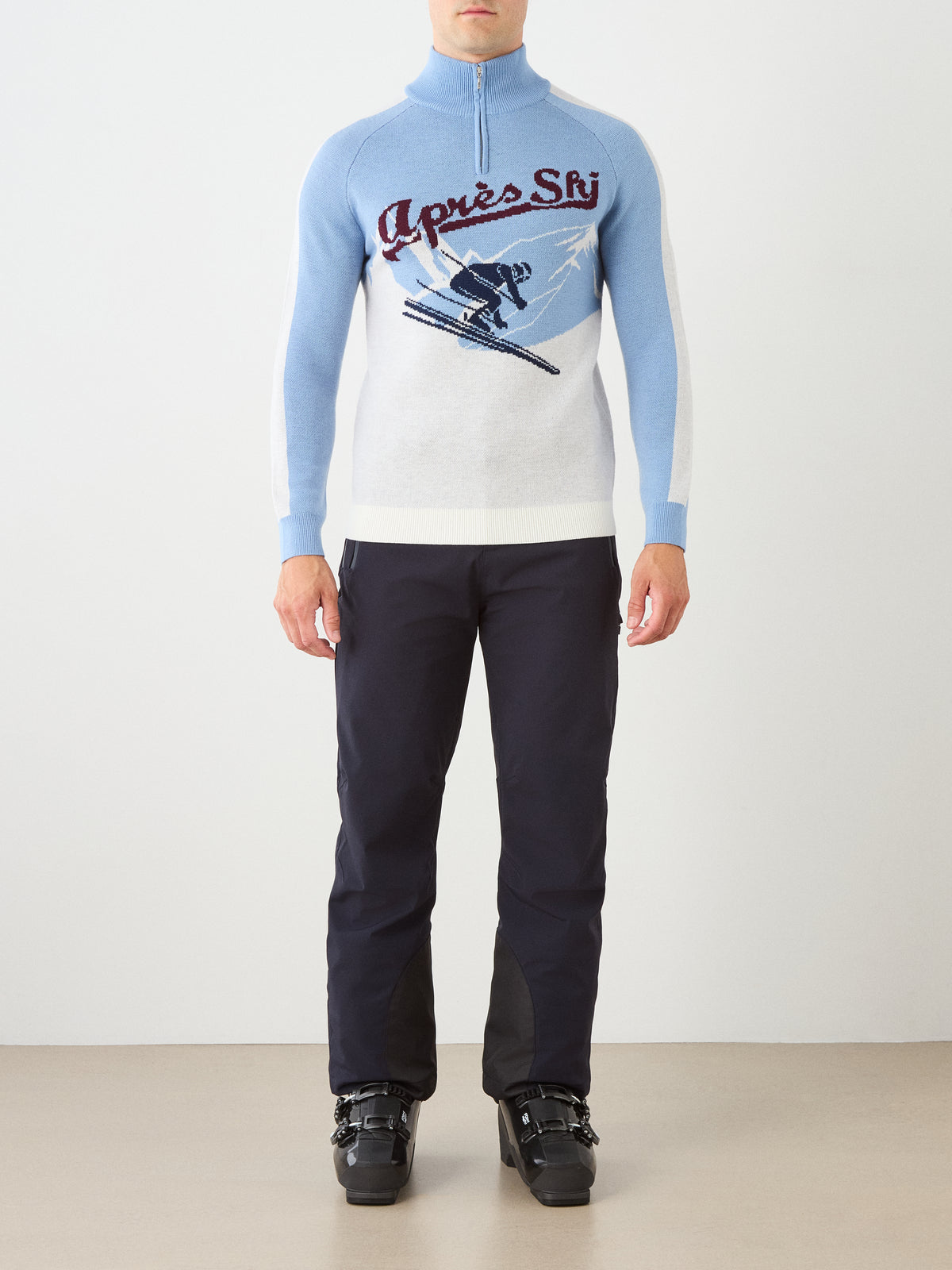 A person stands indoors facing forward against a plain background, wearing the We Norwegians Afterski Sweater Men—a blue and white sweater inspired by vintage ski styles with a charming skier graphic—paired with black pants and black ski boots.