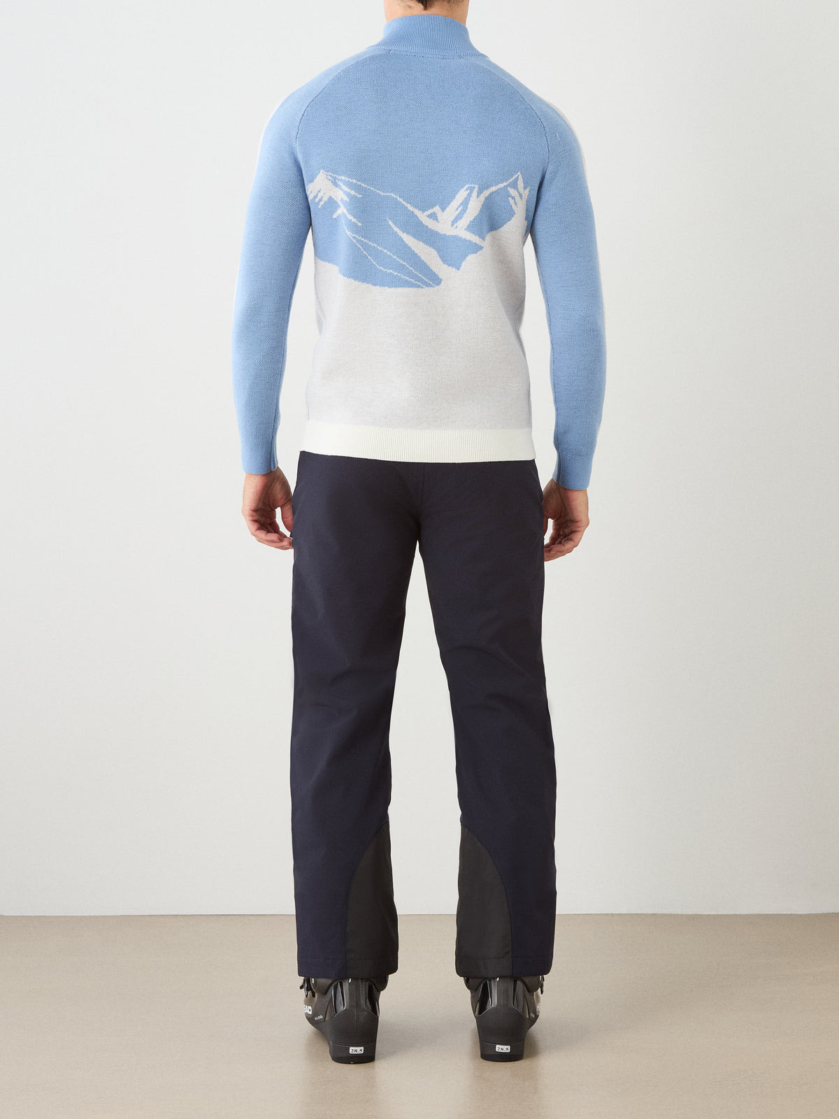 A person stands facing away, wearing the We Norwegians Afterski Sweater Men—a blue and white vintage ski sweater featuring a mountain design on the back—paired with dark pants and black boots. The background is plain and light-colored.