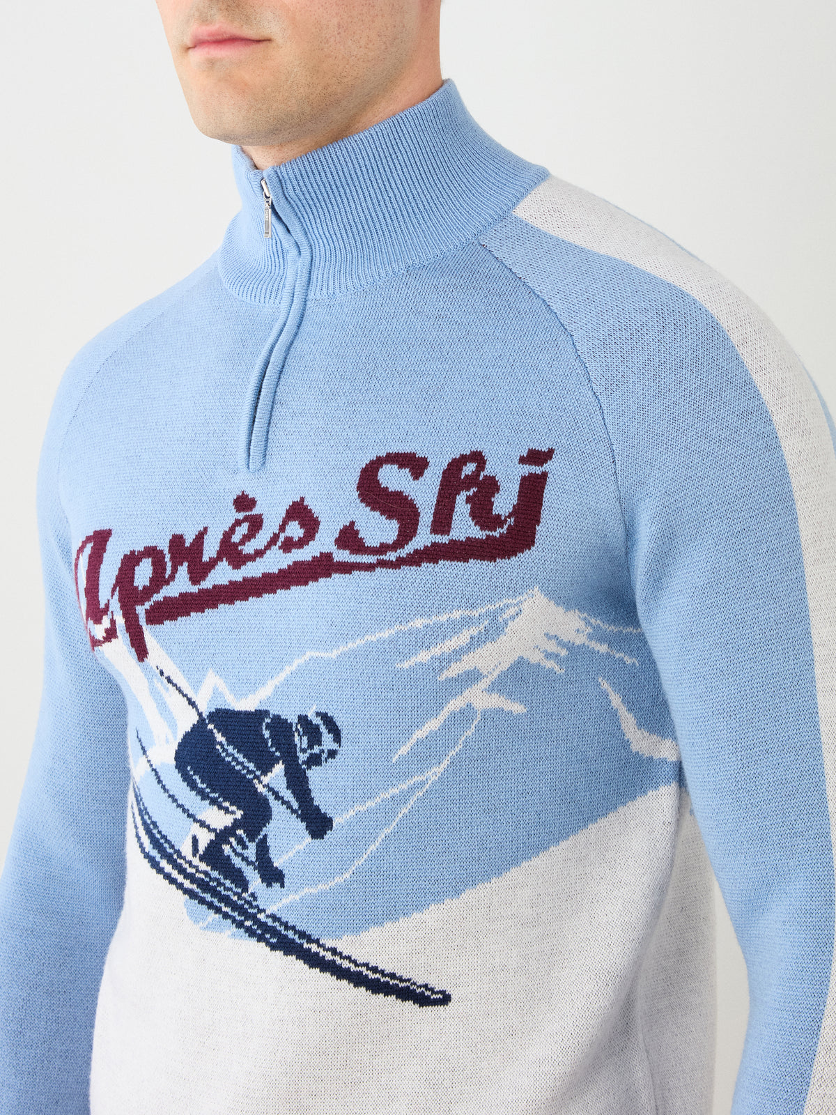 A person wearing the We Norwegians Afterski Sweater Men, a blue and white vintage-style ski sweater featuring “Après Ski” text and a skier graphic set against snowy mountains; only the lower face and upper body are visible.