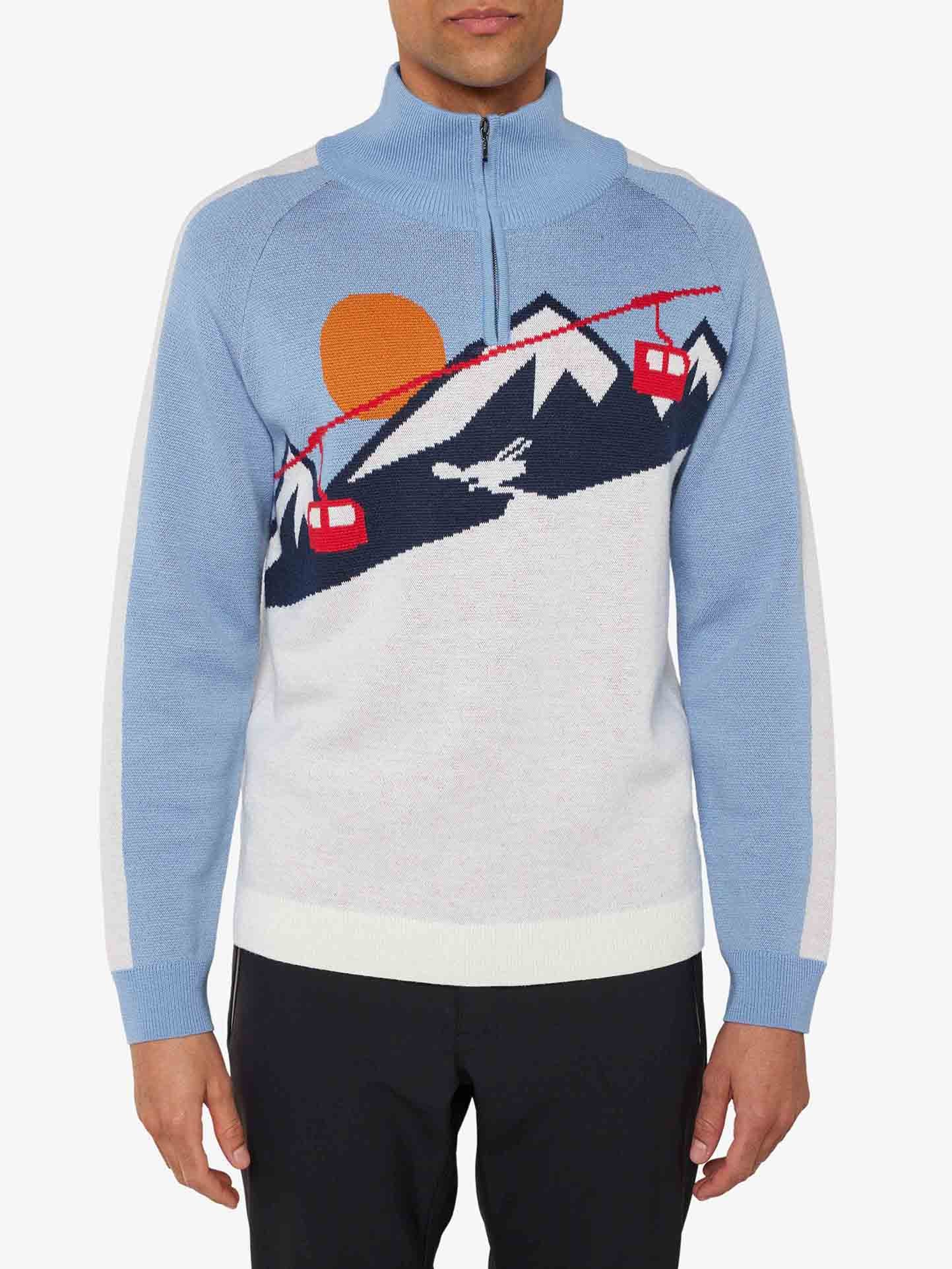 A man is sporting the We Norwegians Afterski Sweater Men in light blue, featuring white side panels and a nostalgic ski design with mountain scenes, a red sun, red cable cars, and a skier. This high-neckline sweater includes a zipper and offers the convenience of washable merino wool. He pairs it seamlessly with black pants.