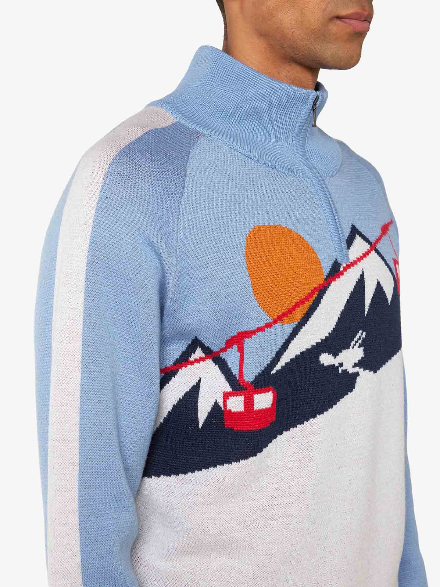 A person is wearing the Afterski Sweater Men by We Norwegians. This blue and white knit sweater with a quarter-zip collar features a retro ski design, showcasing a mountain scene with red ski lift cables, a red cable car, a skier, and an orange sun. Made from washable merino wool, it's perfect for the après-ski vibe.