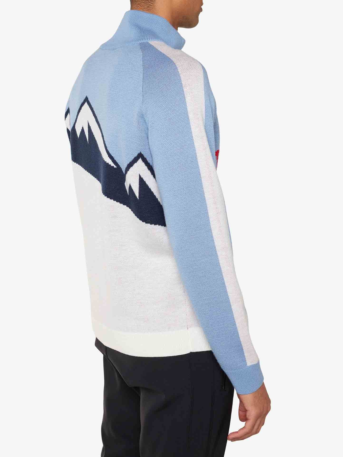 A person wearing the We Norwegians Afterski Sweater Men in light blue, white, and black with a retro ski look featuring a mountain design on the back. The view is from a side angle, showcasing the upper body. The sweater has a light blue stripe running down the arms, and black pants are also visible—perfect for an après-ski outing.