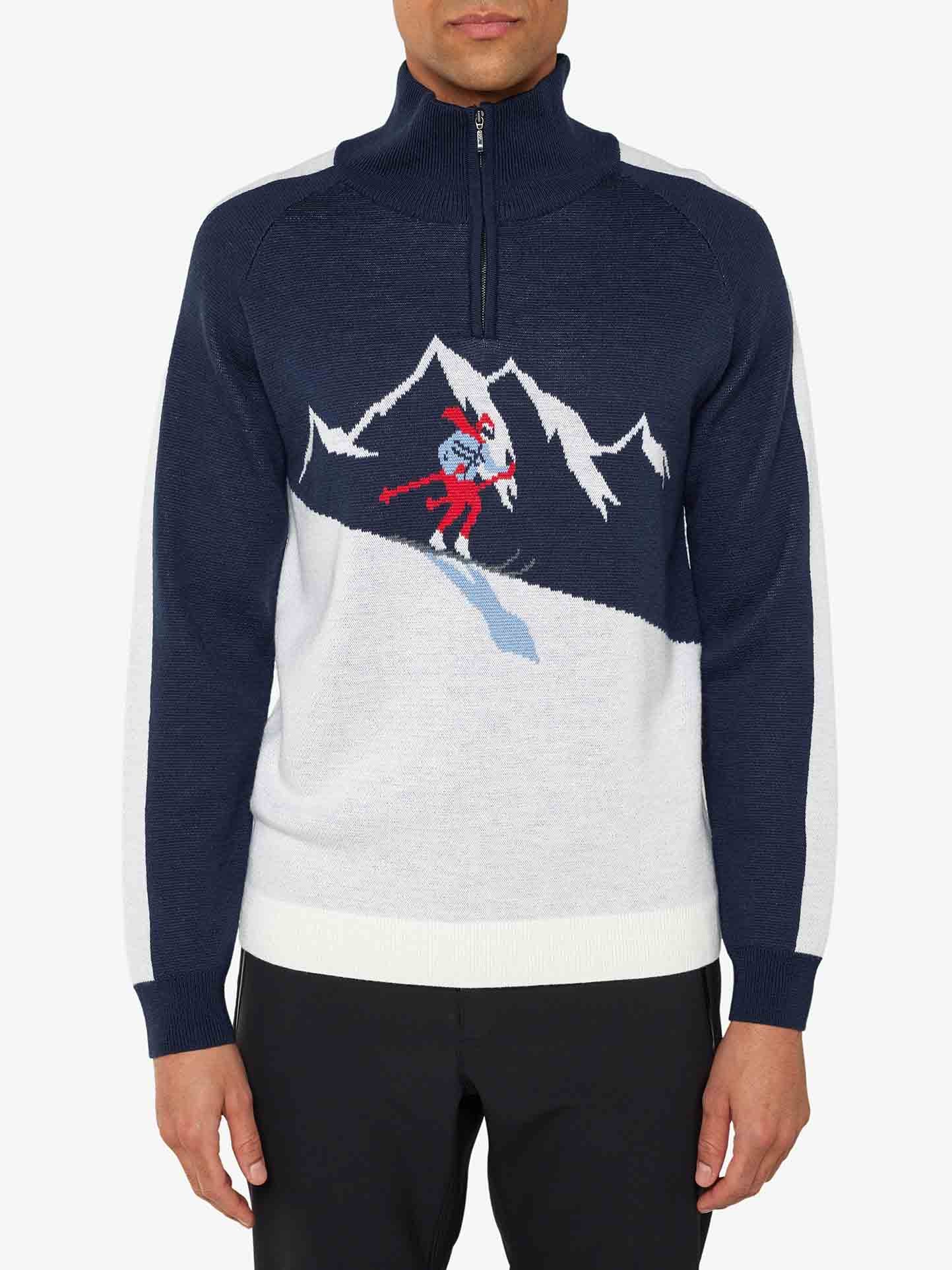 A man wearing the We Norwegians Afterski Sweater Men, a navy blue and white knit sweater with a half-zip collar. This washable merino wool sweater showcases a retro ski look with its design of mountains and a skiing cartoon character in red, white, and blue colors across the chest. He is also wearing black pants.