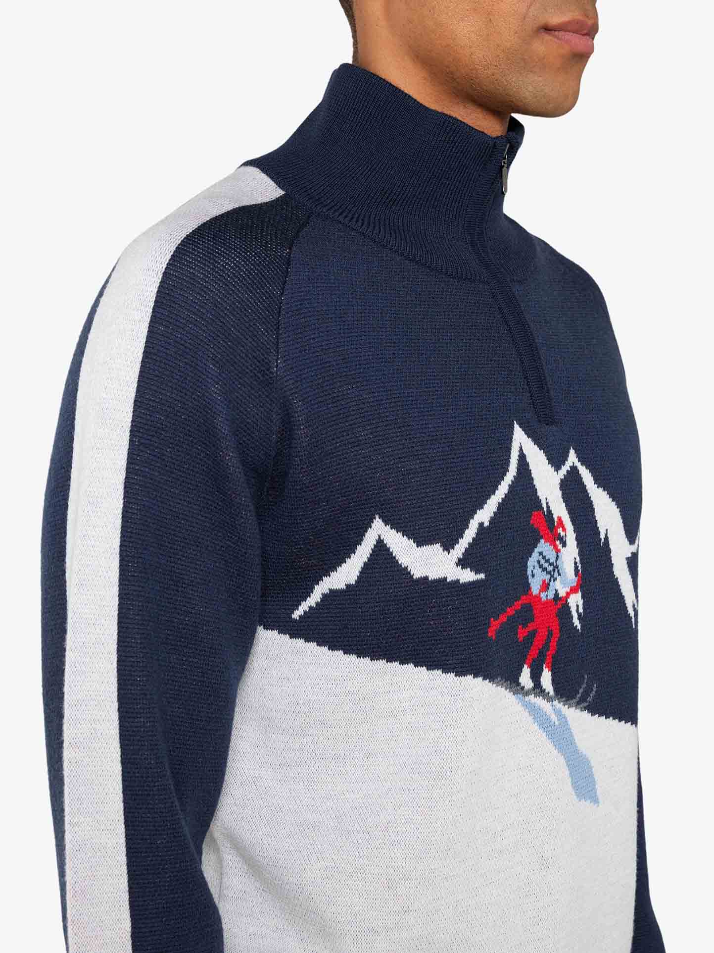 A person is wearing the Afterski Sweater Men by We Norwegians, a dark blue and white zip-neck sweater featuring a skier and mountain design. Made from washable merino wool, it boasts white stripes on the sleeves and a high collar, exuding a retro ski look. The person is standing side-on, facing right.