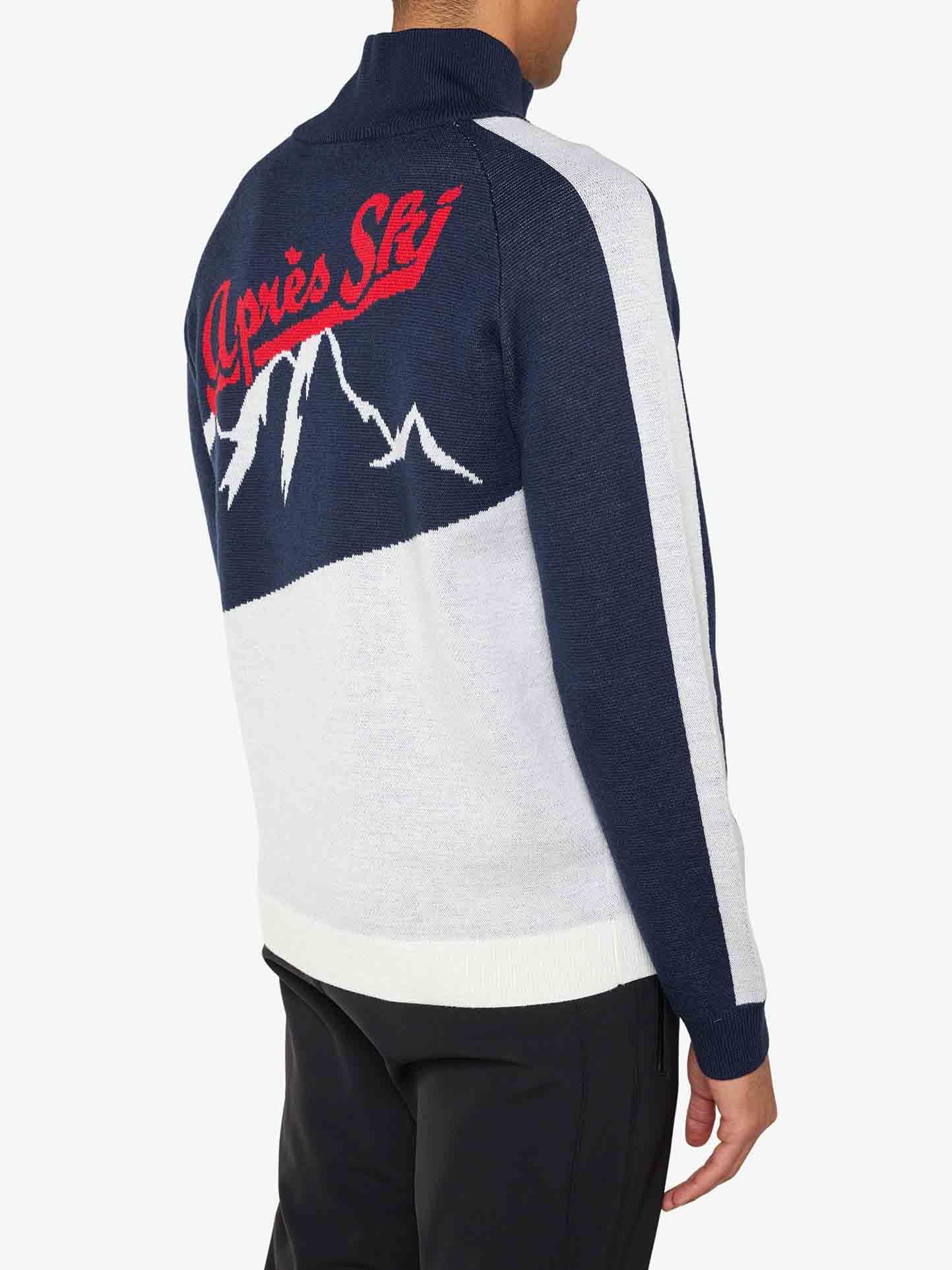 A person is turned away from the camera, showcasing the We Norwegians Afterski Sweater Men, which features a navy blue, white, and red color scheme. Made from washable merino wool, the back of this sweater displays a mountain design along with the words "Après Ski" in red cursive for a retro ski vibe. The individual complements their outfit with black pants.