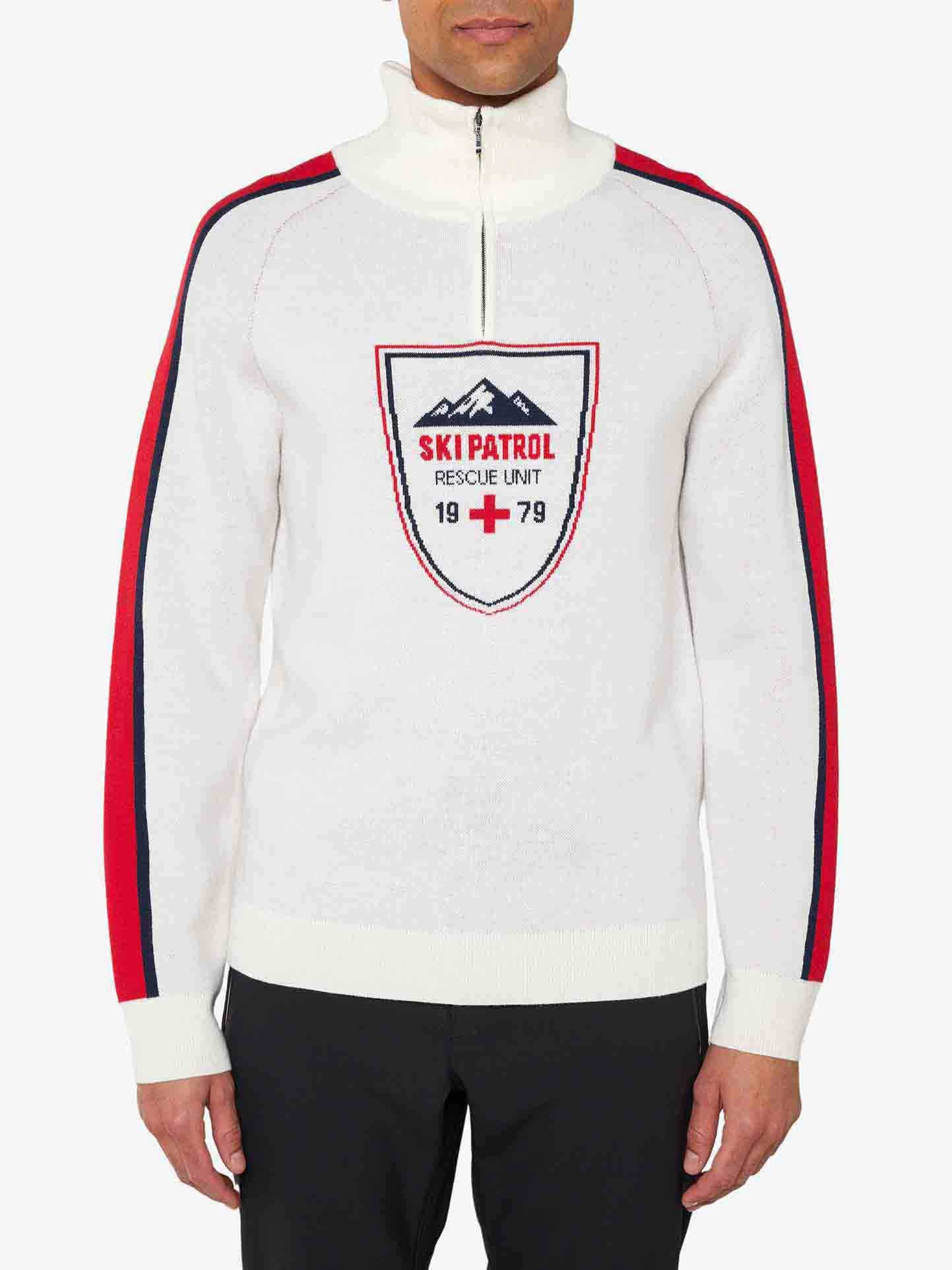 A person stands wearing the We Norwegians' Afterski Sweater Men, a retro ski look white knit sweater with red and blue accents on the sleeves. The sweater features a zip collar, washable merino wool, and a patch on the front with the text "SKI PATROL RESCUE UNIT 1979" along with a red cross emblem. The person is also wearing black pants.