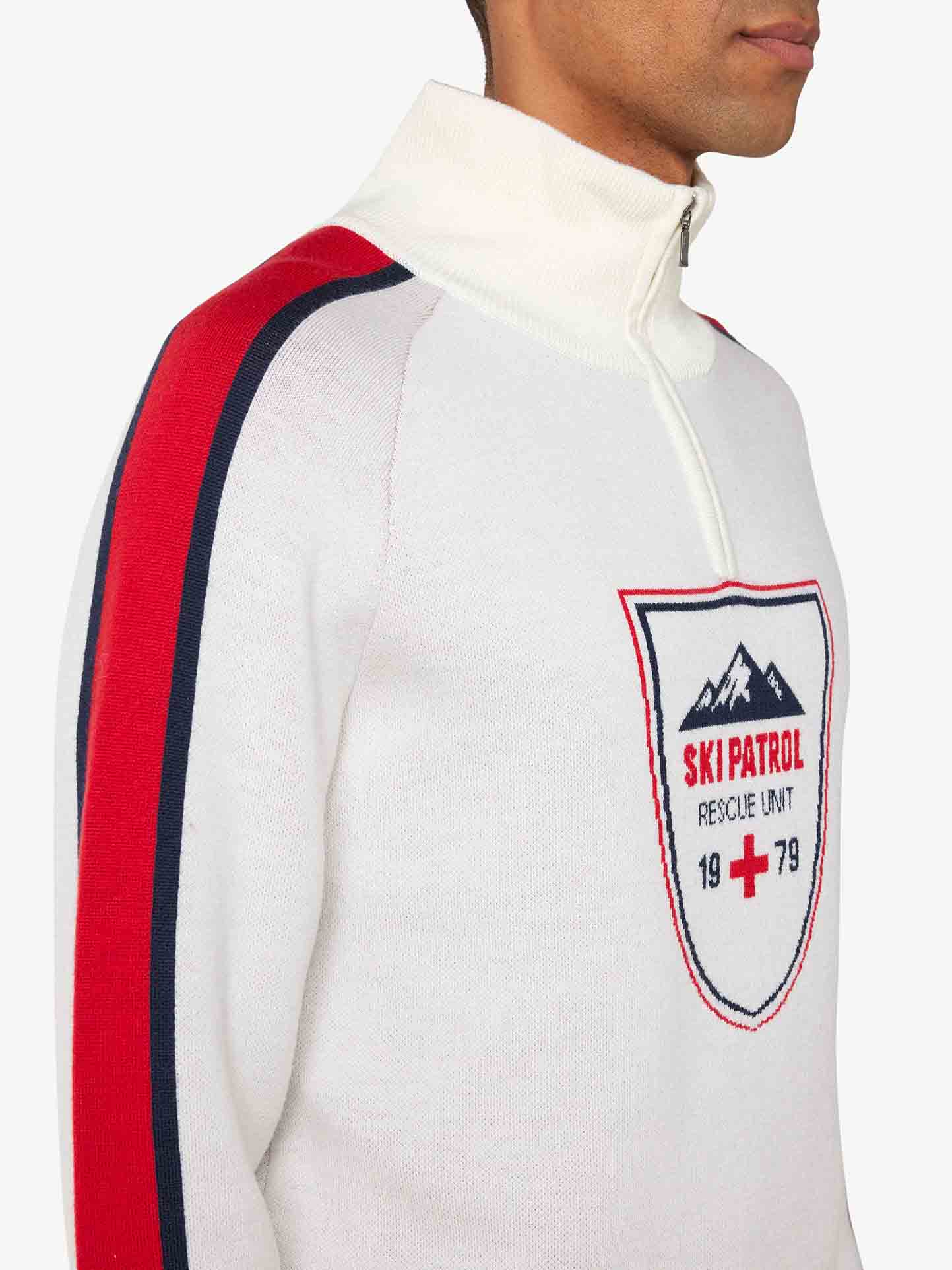 A person wears the "Afterski Sweater Men" by We Norwegians, a white zip-up with a high collar. This retro ski look sweater features red and black stripes on the sleeves and a "Ski Patrol Rescue Unit" badge with mountains, a red cross, and the year 1979 on the chest. Made from washable merino wool, it’s perfect for après-ski moments. The background is plain white.
