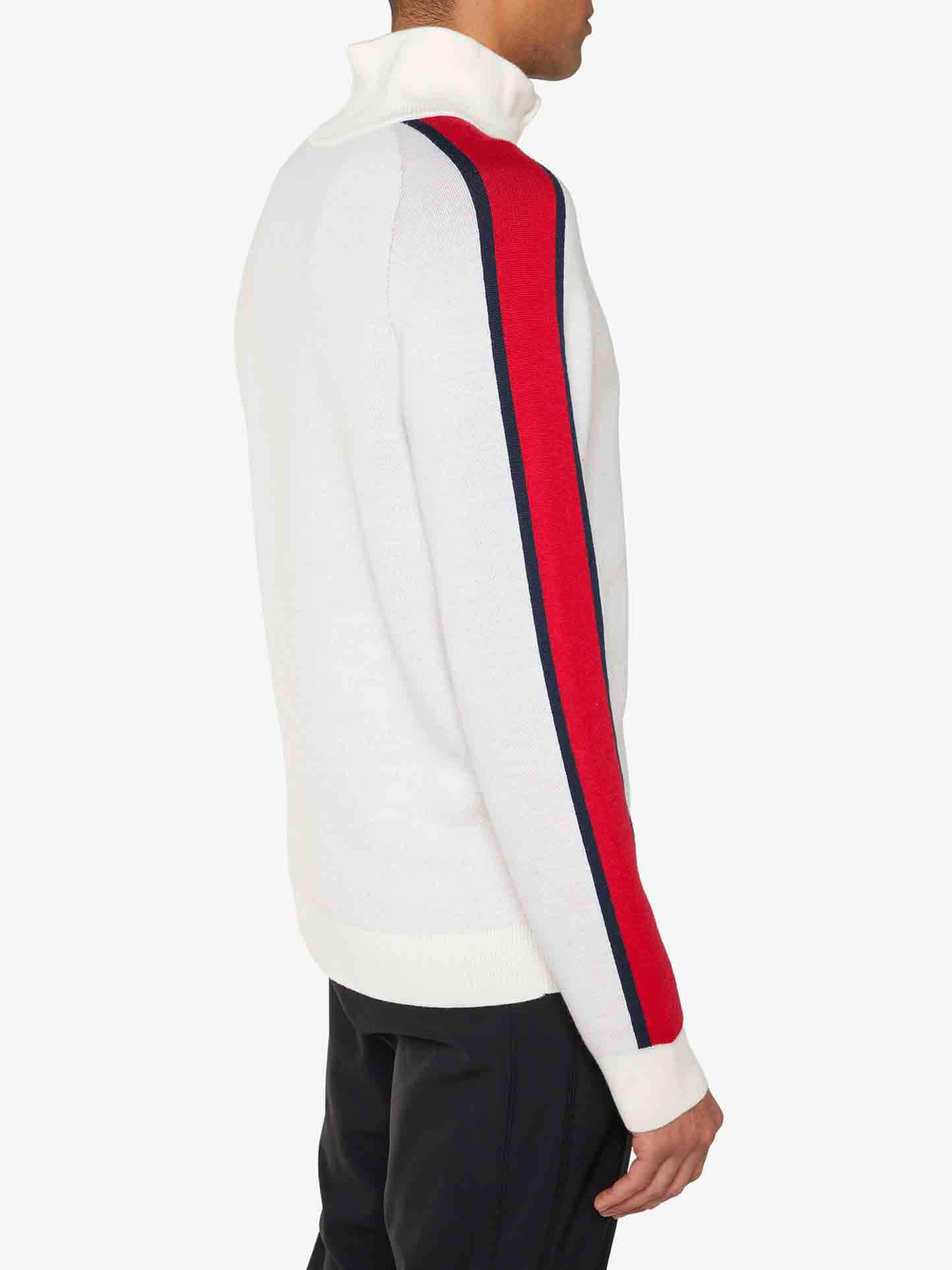 A person is seen standing with their side facing forward, showcasing the back of a We Norwegians Afterski Sweater Men, which features red and navy blue stripes running down the sleeves of its white turtleneck design. The retro ski ensemble is completed with black pants.