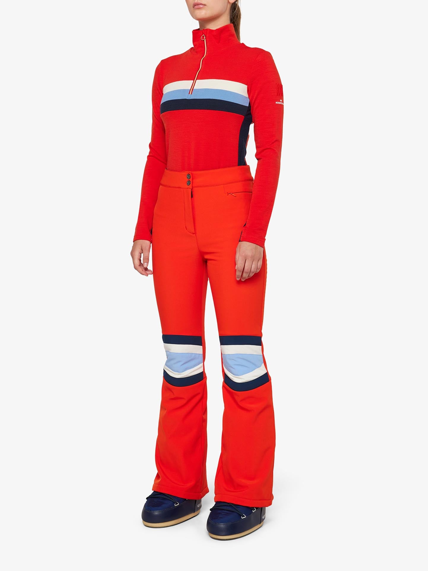 A woman stands wearing a vibrant red ski outfit, featuring a long-sleeve top with black and white stripes and high-waisted We Norwegians Snowfun Ski Pant Women with blue and white panels on the thighs. She also sports black ski boots. The background is plain white.