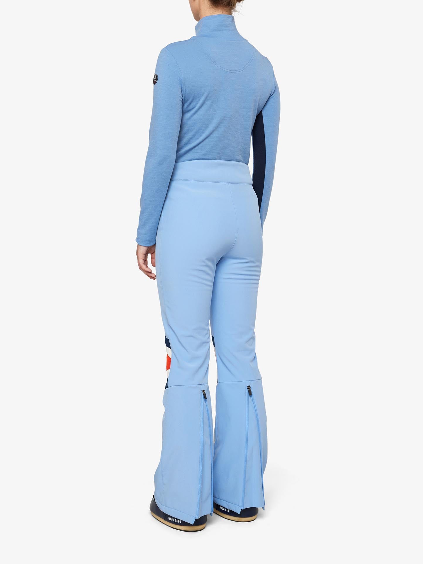 A person stands facing away from the camera, clad in a blue long-sleeve top and matching We Norwegians Snowfun Ski Pant Women with zippered bottoms. The form-fitting outfit is made from stretchy soft shell material, making it ideal for cold weather activities like skiing or other snow-related fun.