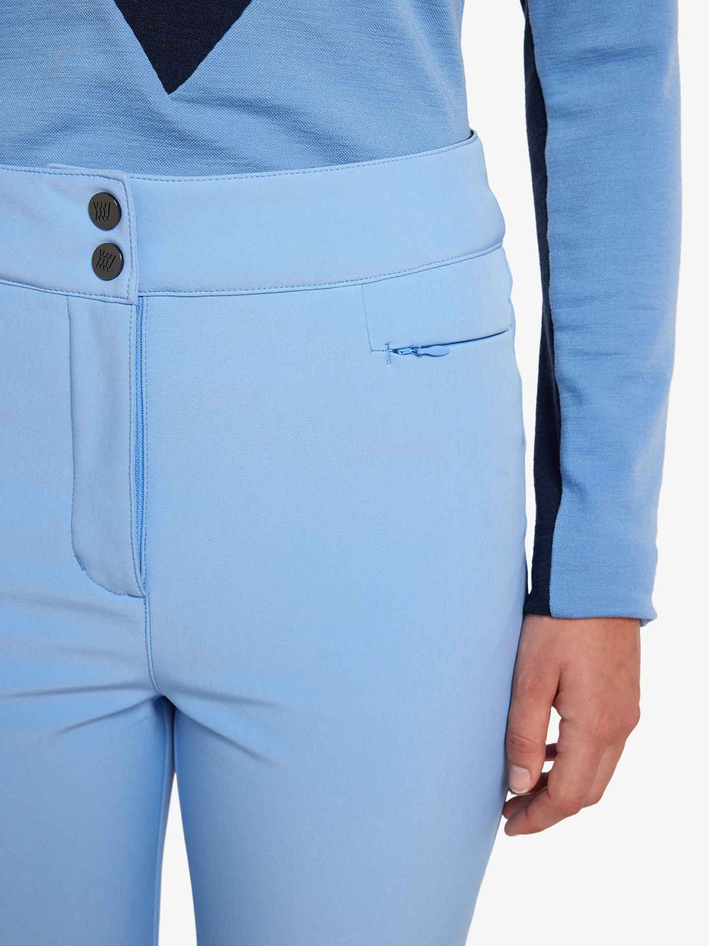 A close-up of a person wearing We Norwegians' light blue Snowfun Ski Pant Women, with a partial view of a light blue and dark blue long-sleeved shirt. The ski pants feature two front buttons and a small zippered pocket on the right side. The person's right hand is visible resting to the side.