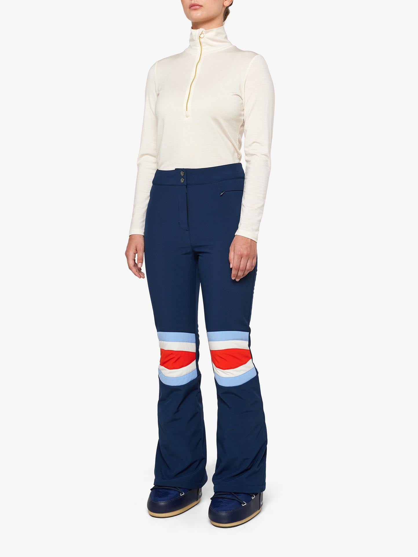 A person is wearing a light beige long-sleeve turtleneck top and navy blue Snowfun Ski Pant Women by We Norwegians, featuring white, red, and light blue stripes on the knees. They are also wearing dark blue snow boots. The background is plain white.