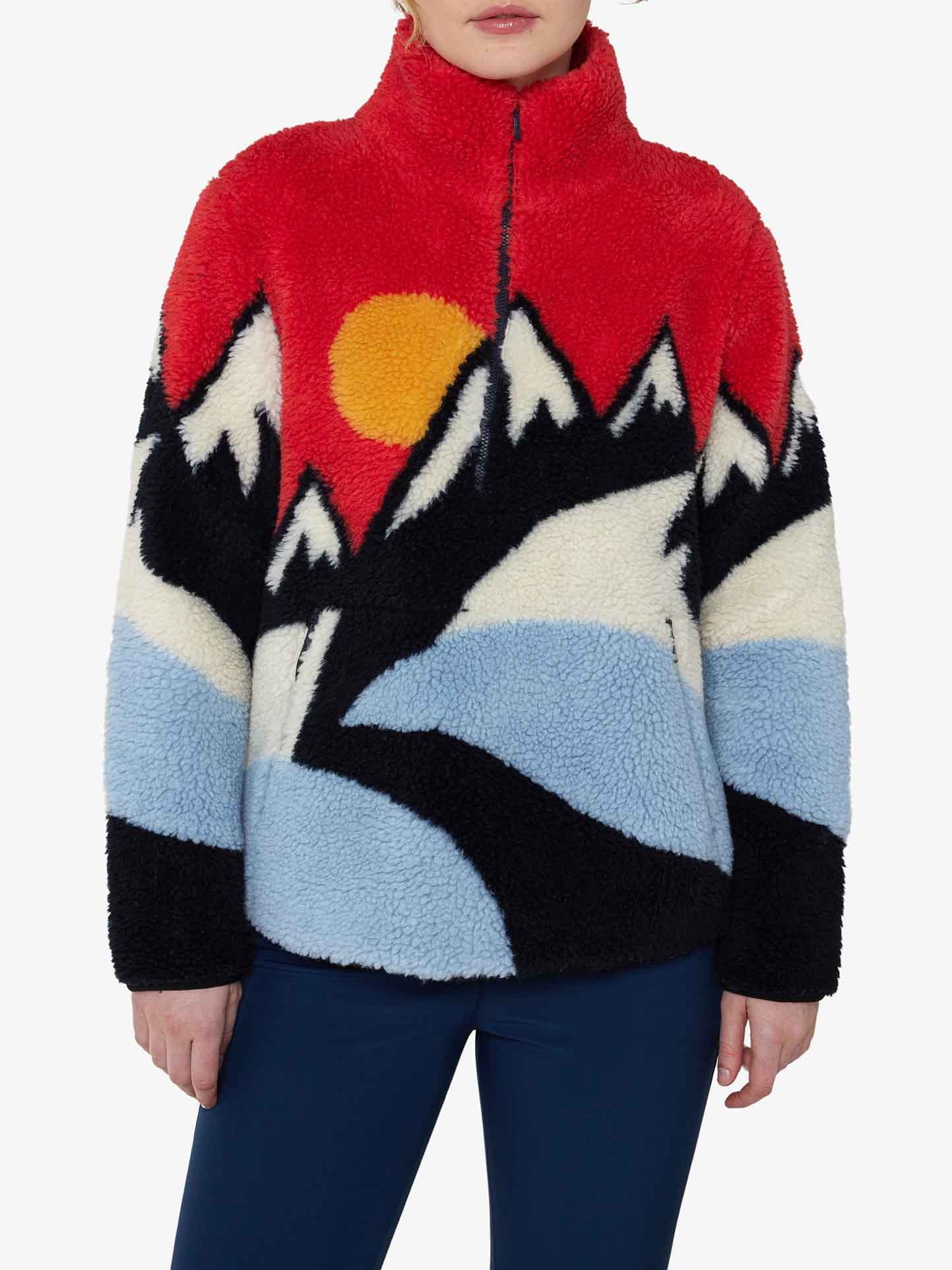 A person wearing the Snowfun Sweater Women by We Norwegians, adorned with a vibrant design of mountains and the sun, stands against a plain white background. This fleece jacket features a blend of reds, blues, blacks, and whites reminiscent of snowy peaks.