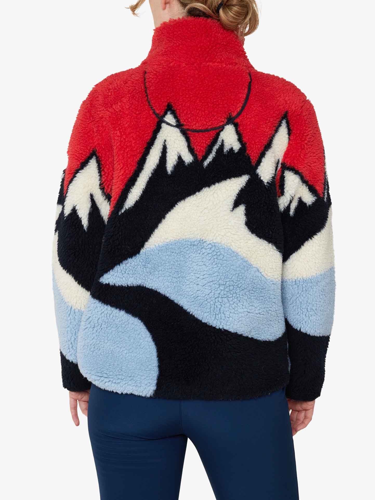 A person is seen from behind wearing the We Norwegians Snowfun Sweater Women, which features vibrant mountain and wave patterns in red, black, white, and blue. The jacket's shearling appearance complements their blue pants perfectly, creating an adventurous vibe reminiscent of snowy mountains.