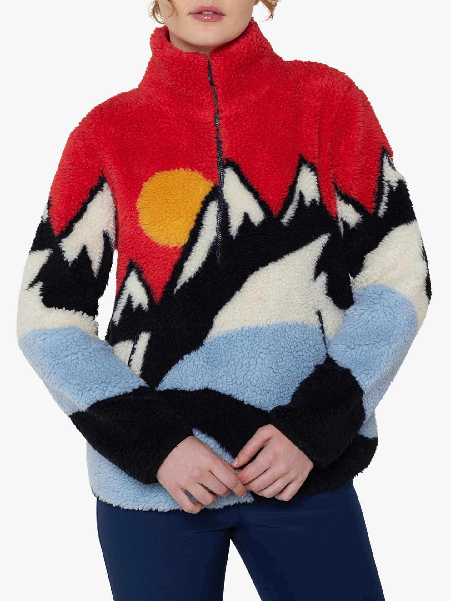 A person is wearing the Snowfun Sweater Women by We Norwegians, showcasing a colorful fleece design with a shearling look. It features a mountain landscape design with a red sky and yellow sun, complemented by black and light blue sleeves. They stand against a plain background.