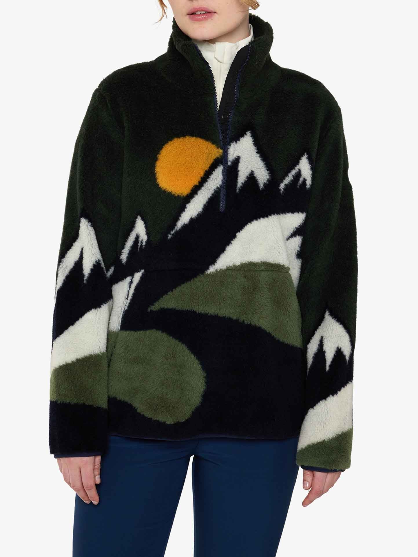 A person is sporting the Snowfun Sweater Women by We Norwegians, a black pullover showcasing snowy mountains and a sun design in white, green, and yellow. Crafted from merino wool, it features a high collar and is complemented by dark blue pants, with the person's face obscured from view.