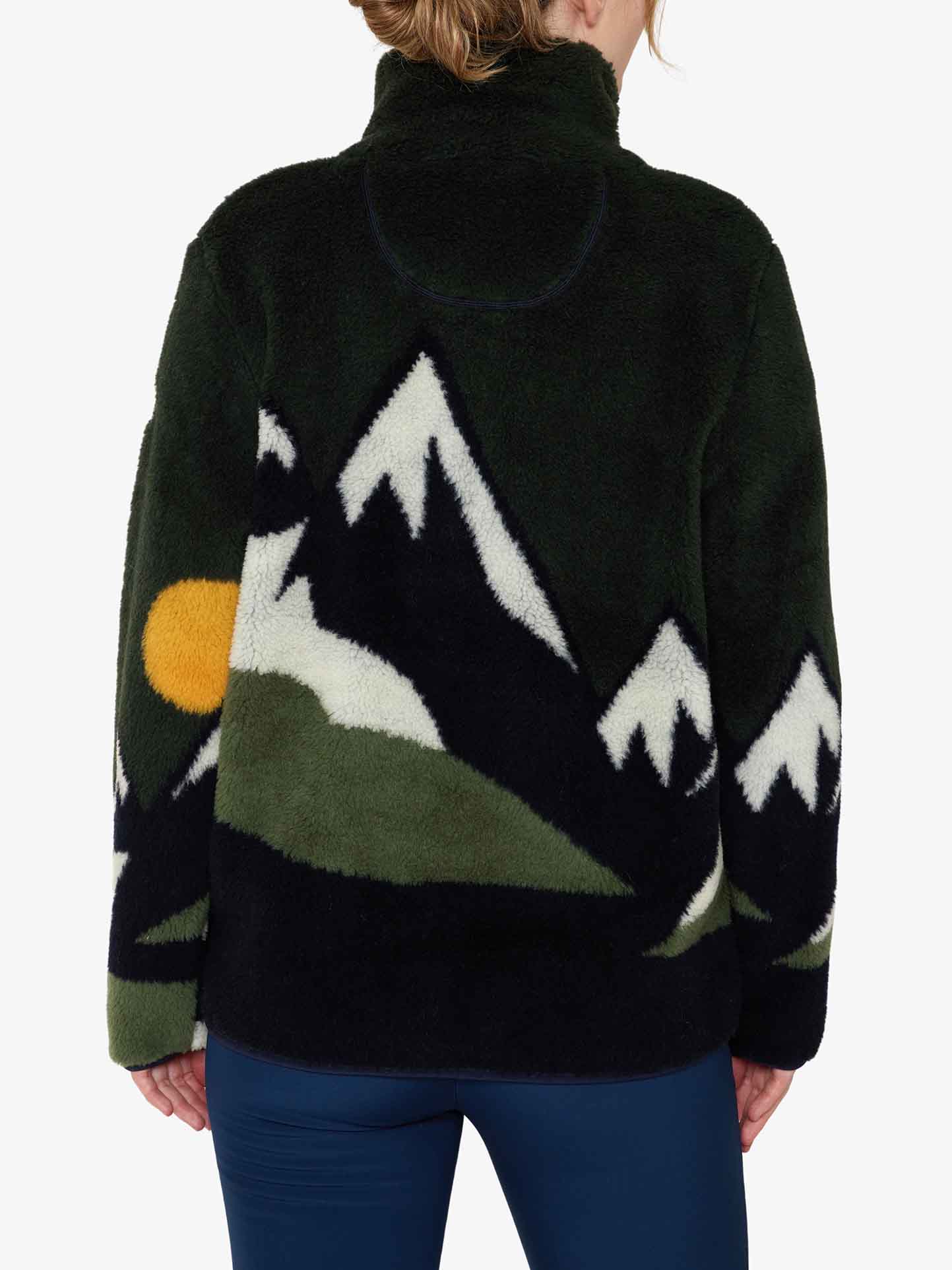 A person wearing the We Norwegians Snowfun Sweater Women, which boasts a fluffy black texture with designs of snowy mountains and a sunset on the back. The shearling-look sweater showcases white peaks, green slopes, and a yellow orb symbolizing the sun. The individual is facing away from the camera, dressed in dark blue pants.