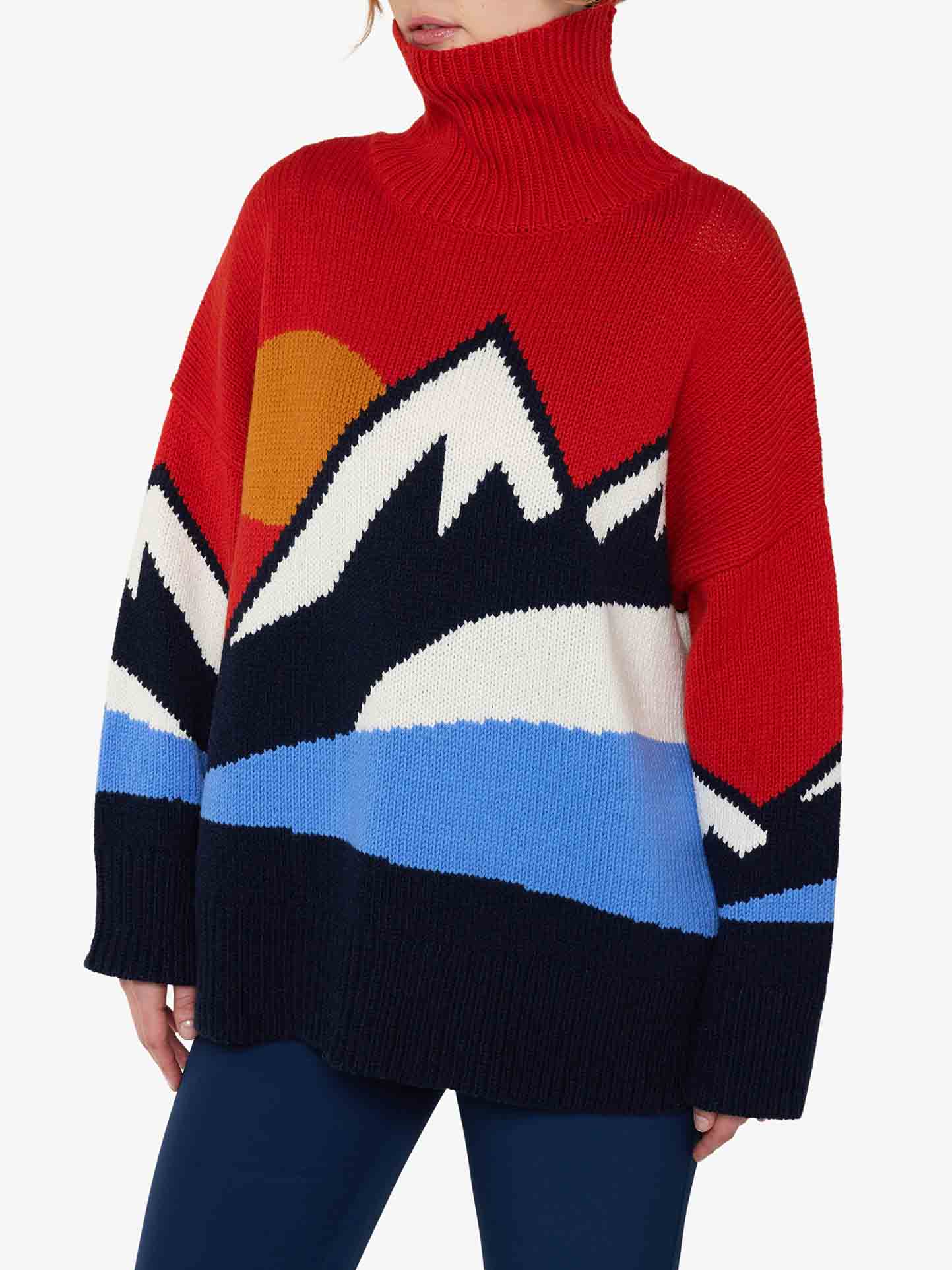 A person is wearing the Snowmountain Sweater Women by We Norwegians, a cozy red turtleneck featuring a colorful mountain pattern in black, white, blue, and yellow that extends across the chest and sleeves, perfectly embodying Fall fashion vibes. They are also dressed in dark blue pants. The head is partially cropped out of the image.