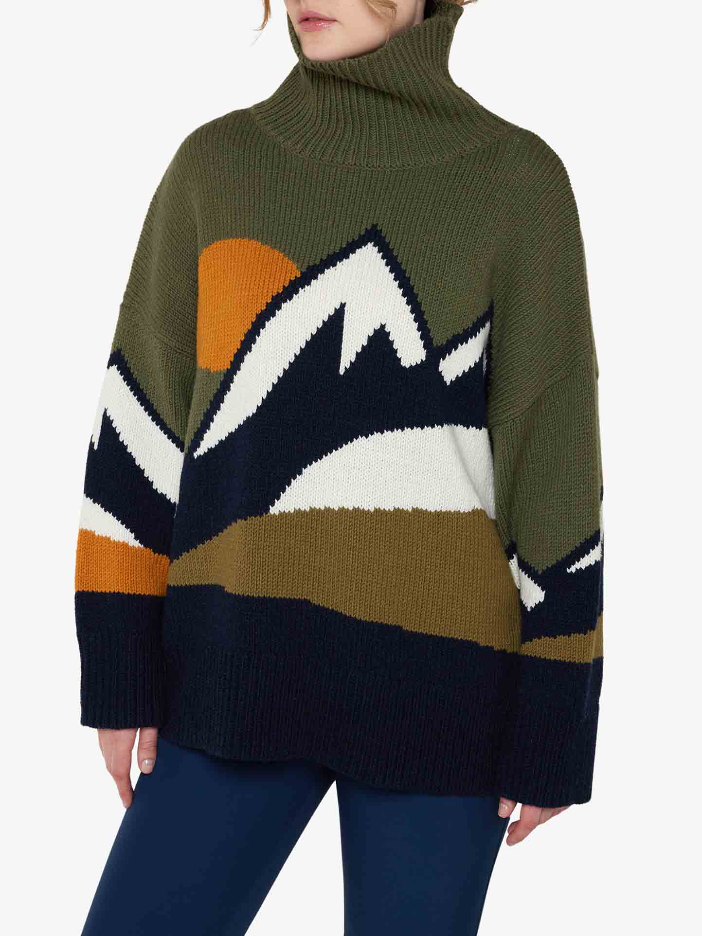 A person is wearing a loose-fitting, olive-green Snowmountain Sweater Women by We Norwegians, crafted from cozy Merino wool and adorned with a mountain landscape design in navy, white, orange, and brown. Paired with dark blue pants against a plain white background, this look encapsulates the essence of fall fashion perfectly.