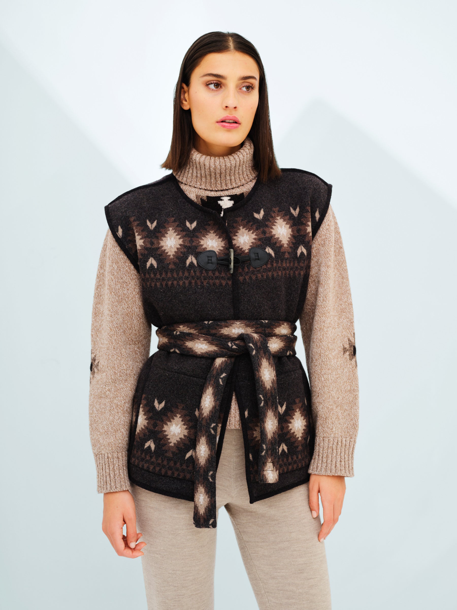 A woman with straight, shoulder-length brown hair is wearing a beige turtleneck sweater layered under the dark, patterned Hovden Vest Women by We Norwegians. The vest is belted at the waist. She stands against a light background, looking away from the camera with a neutral expression.