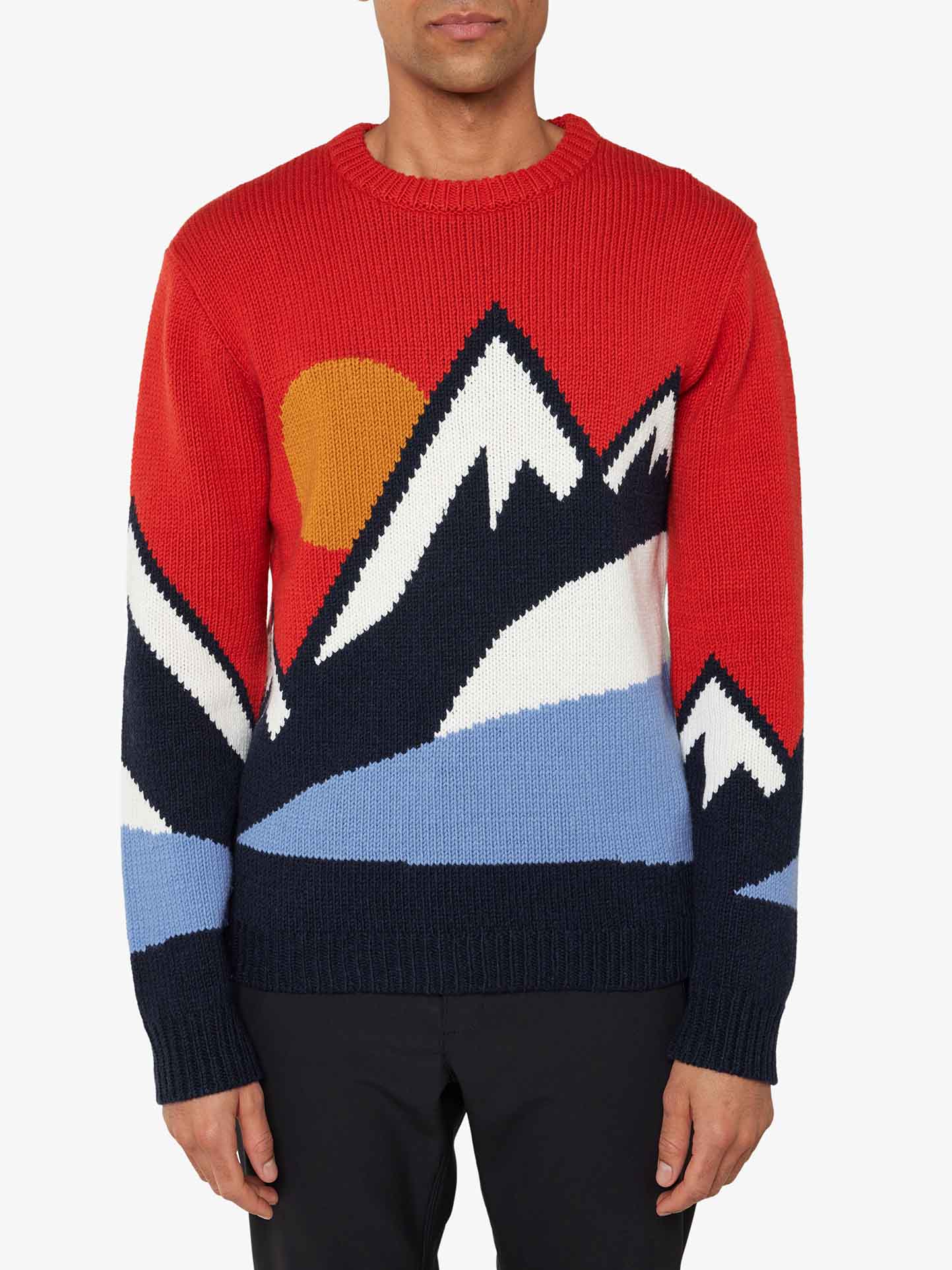 A person wearing the We Norwegians Snowmountain Sweater Men, a cozy red knitted garment featuring a stylized mountain landscape design with snow-capped peaks, a sun setting behind the mountains, and contrasting colors of blue, white, and black. The merino wool blend adds extra warmth. The person is shown from the chest down.