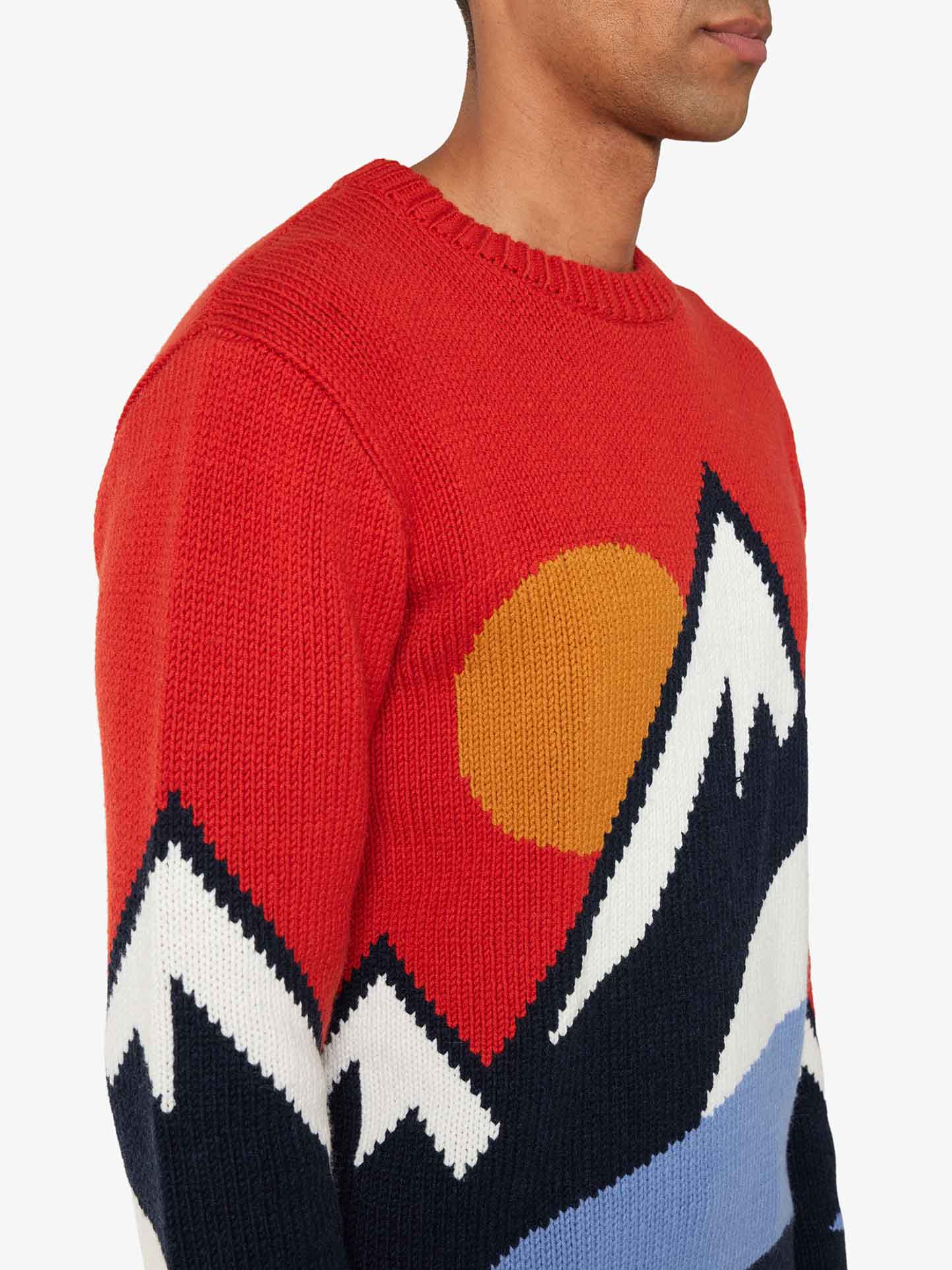 A person wearing the Snowmountain Sweater Men by We Norwegians, showcasing a vibrant and cozy design with a mountain range featuring white peaks, dark valleys, a red upper background, and an orange sun. The luxurious cashmere mix adds to the opulence as the person is viewed from the side, highlighting the colorful and bold pattern of the sweater.